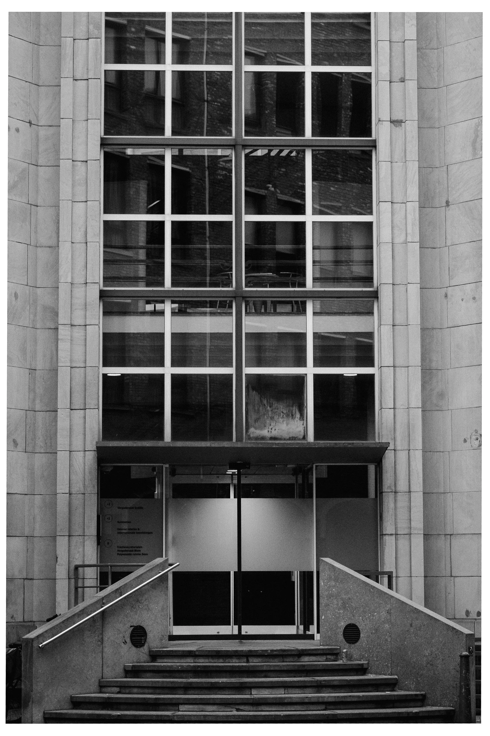 Voorgevel van een modern gebouw met grote glazen ramen, toegang via trappen, en een metalen deur.