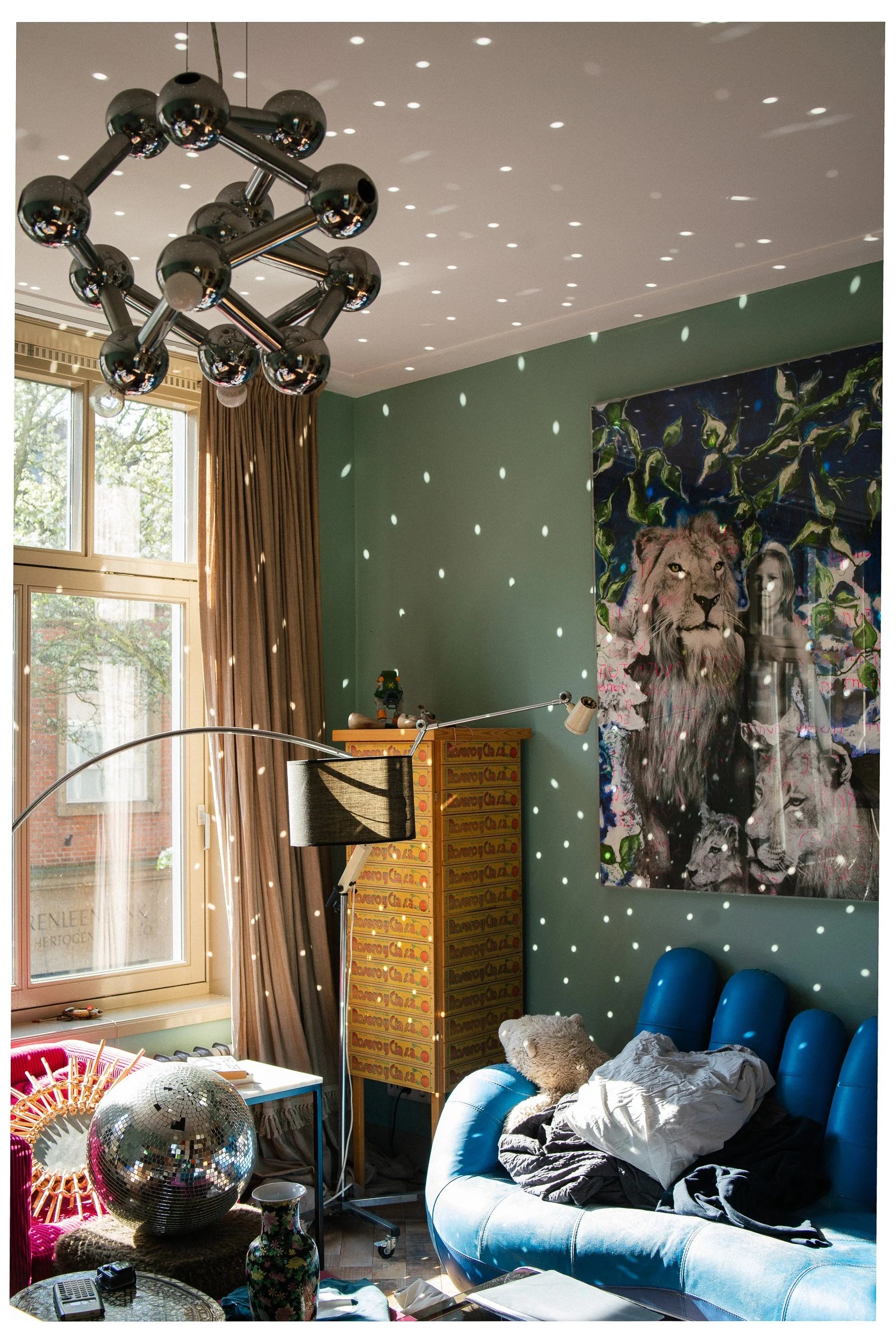 Een kleurrijke woonkamer met een groene muur, een groot raam met goudkleurige gordijnen, een blauwe bank met een teddybeer en kleren erop, een gouden stolp en een disco bal, een lamp met een lange arm, een kast met gele laden en een grote muurkunstwerk met een leeuw en een vrouw.