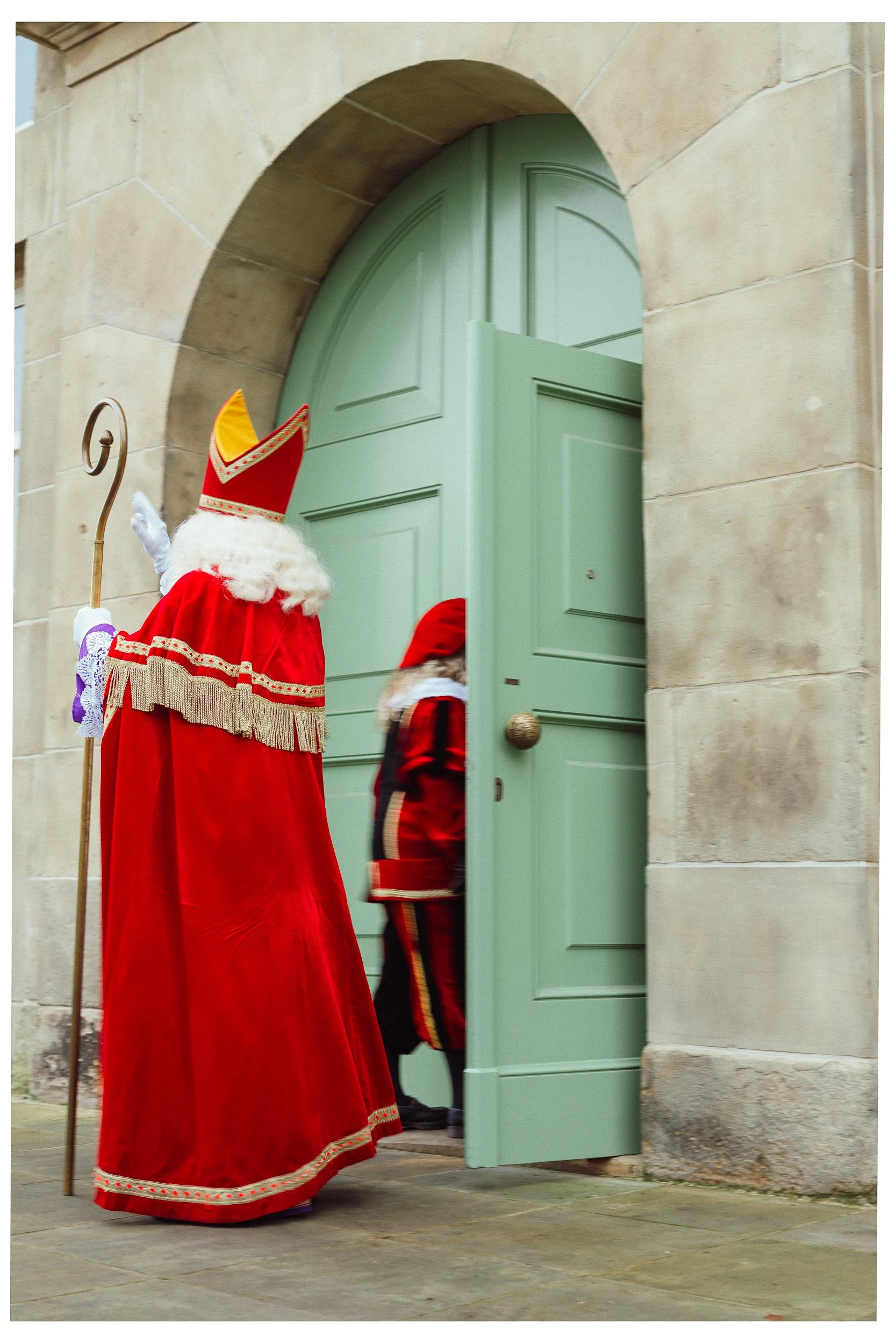 Sinterklaas en Zwarte Piet bij een groene deur, Sinterklaas draagt een rode mantel en mijter, Zwarte Piet draagt een rood en zwart kostuum.