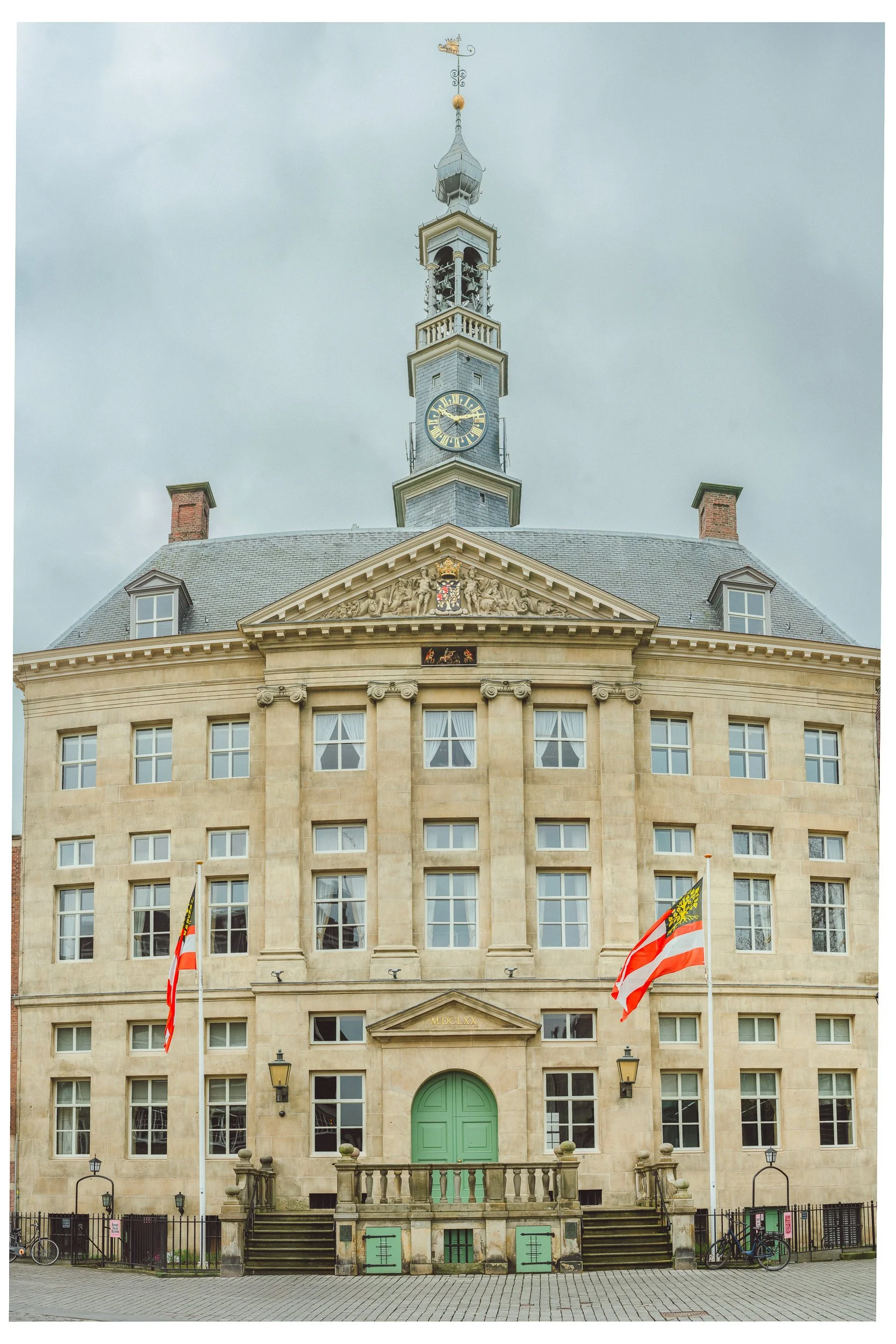 Een imposinggebouw met een klok aan de top, vlaggen voor de ingang, en een paardenkoets op de voorgrond, onder een bewolkte lucht.
