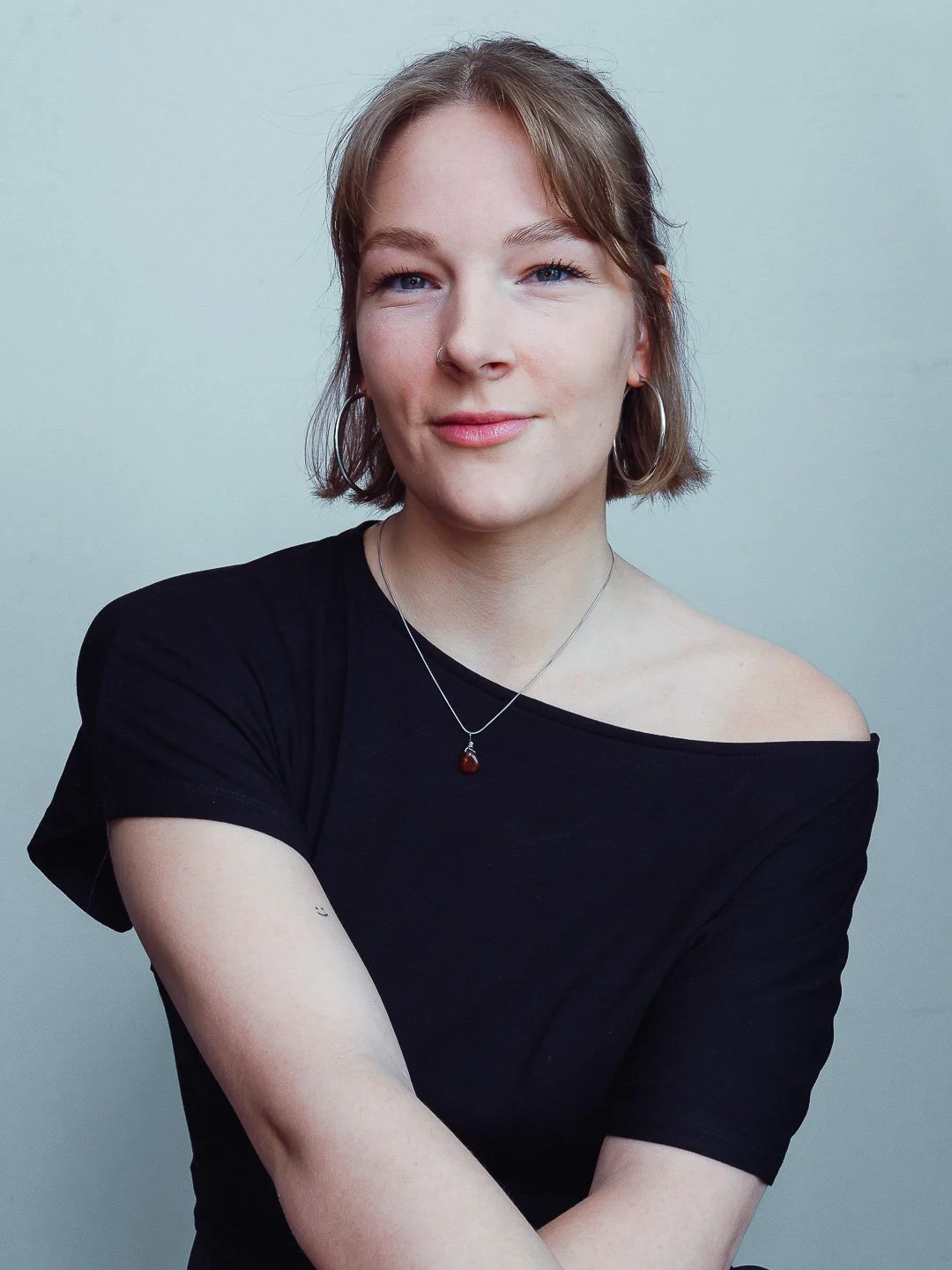 Portret van een vrouw met kort, lichtbruin haar en blauwe ogen, die een zwarte off-shoulder top draagt en een ketting met een rood hanger draagt.