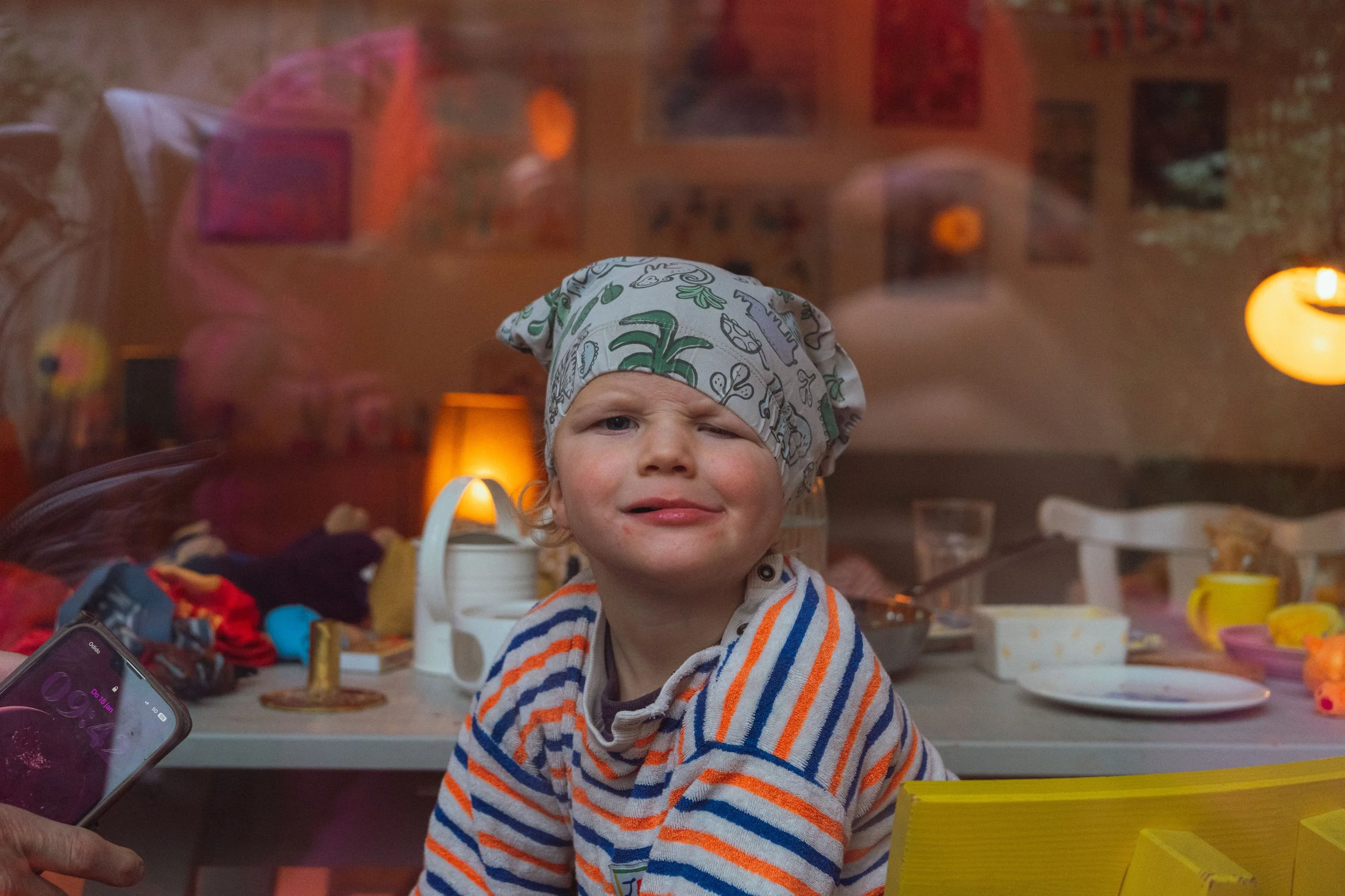 Een jonge jongen met een dierenprint hoed en een gebreide trui met oranje en blauwe strepen zit aan een tafel, gekleed voor een feestje of activiteit.