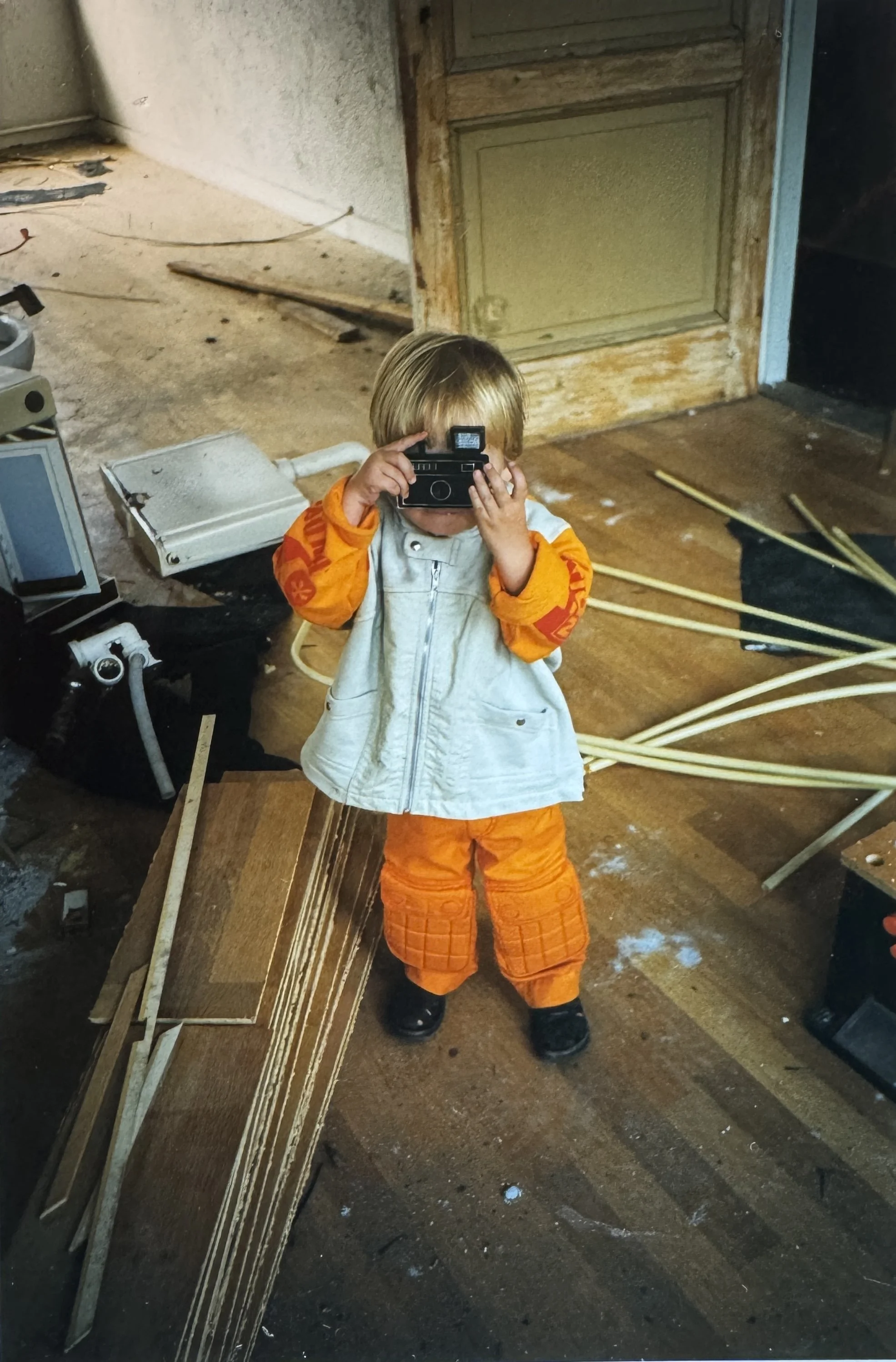 Een jongen met blond haar en een oranje jack met patroon, die een oude camera vasthoudt en omhoog wijst, staat in een rommelige kamer met schroot, draden en vuil op de vloer.