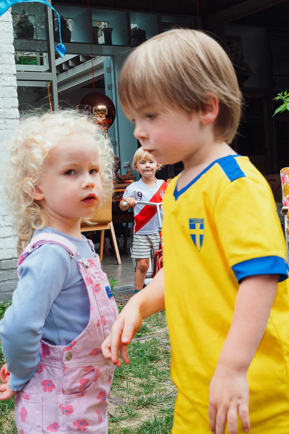 Twee jonge kinderen, een meisje met blond, krullend haar in een broek met bloemenpatroon en een jongen met lichtbruin haar in een gele voetbaloutfit met het Zwedse vlaglogo, staan dicht tegen elkaar in een tuin, terwijl een derde kind met een wit en rood shirt op de achtergrond op een speelgoedfietsje staat.