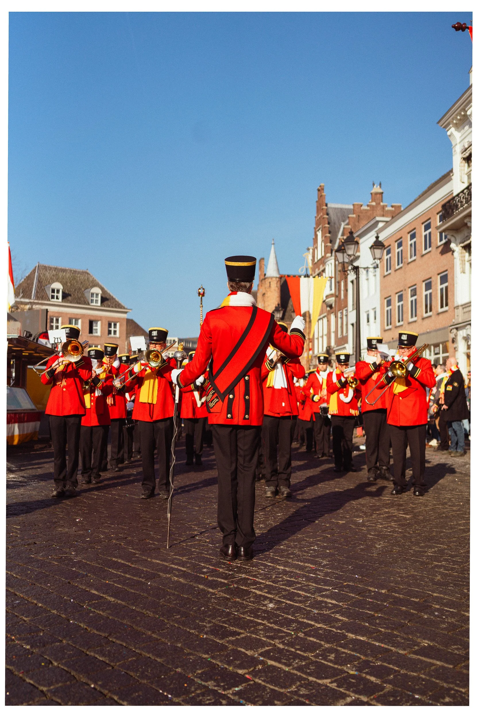 Een muzikale parade met een brassband in rode uniformen en hoeden, optrekkend op een straat met klinkers, onder een blauwe hemel, omlijst door historische gebouwen.