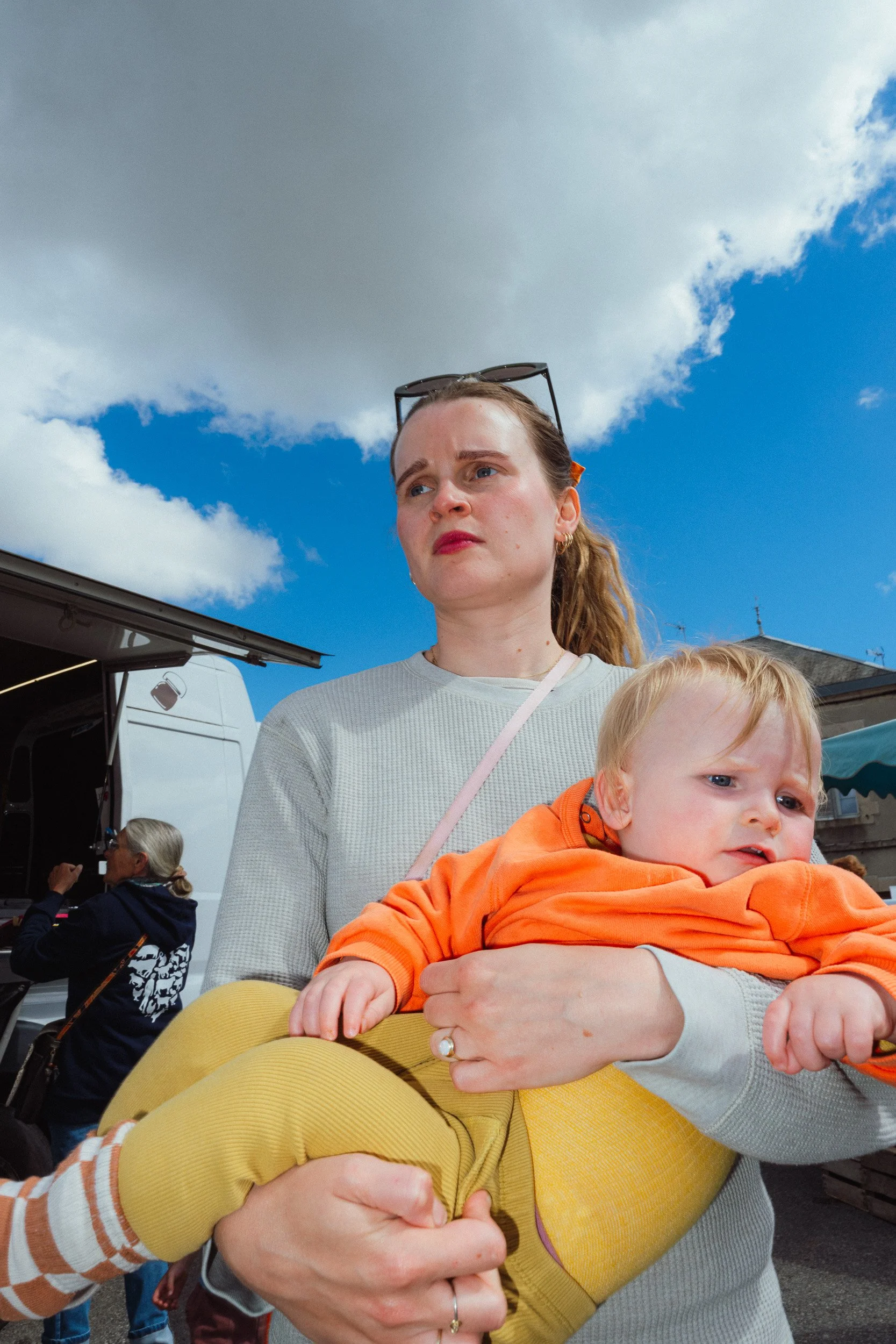 Een vrouw houdt een jonge jongen in haar armen tijdens een buitengebeurtenis, onder een blauwe hemel met wolken.