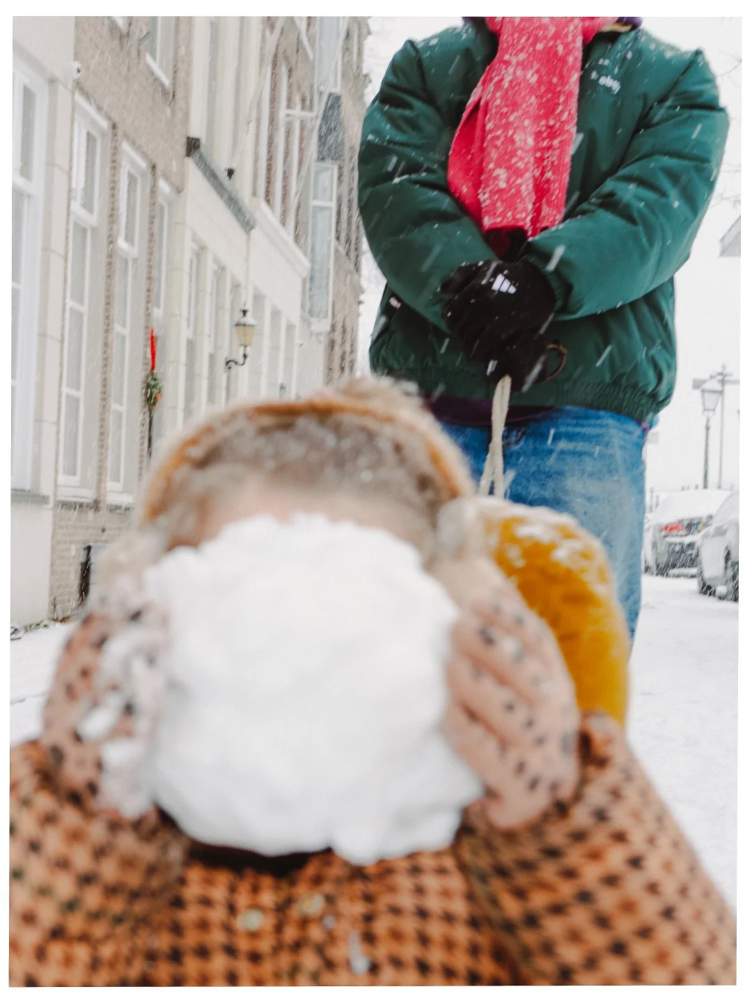 Persoon houdt een warme chocolademelk met slagroom in een sneeuwachtige straat, terwijl een andere persoon op de achtergrond staat in winterkleding.