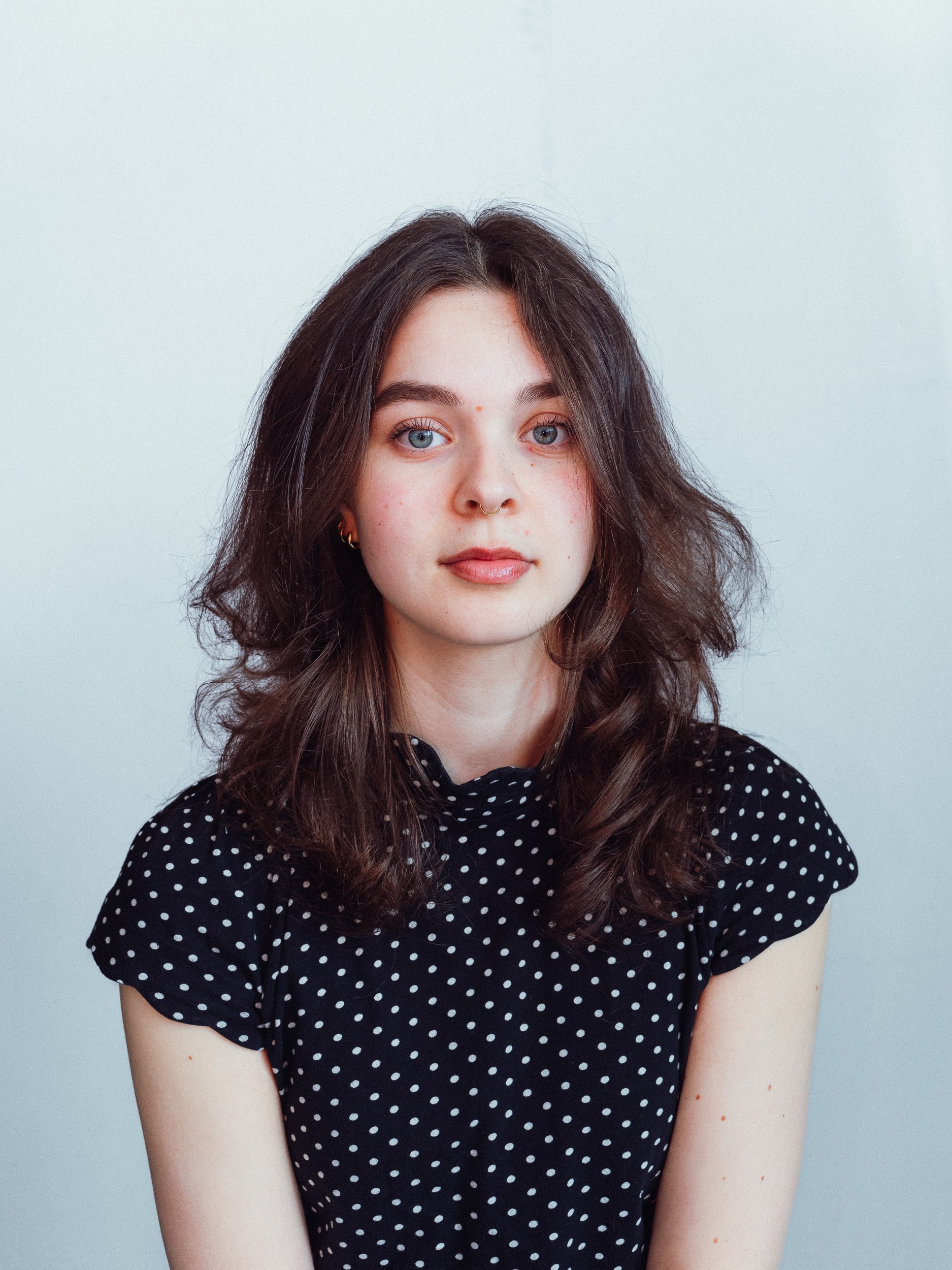 Portret van een jonge vrouw met lang, golvend bruin haar en blauwe ogen, gekleed in een zwarte jurk met witte polkadots, tegen een lichtgrijze achtergrond.