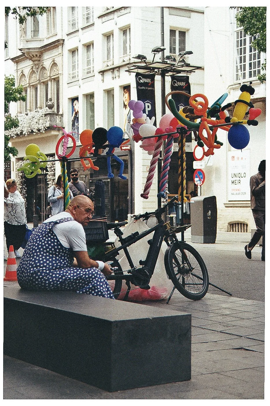 Man in gekleurde stippen pyjama zit op een bankje naast een elektrische fiets, op straat met ballonnen en decoraties voor een evenement.