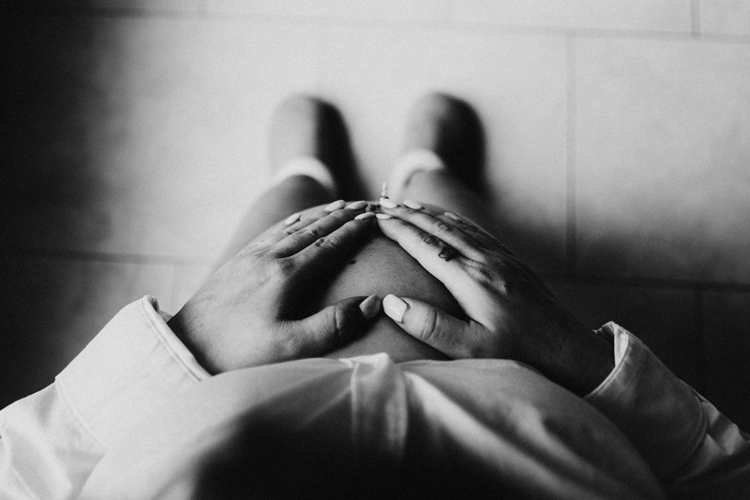 Black and white photo of a person with hands resting on a pregnant belly, wearing a shirt and socks.