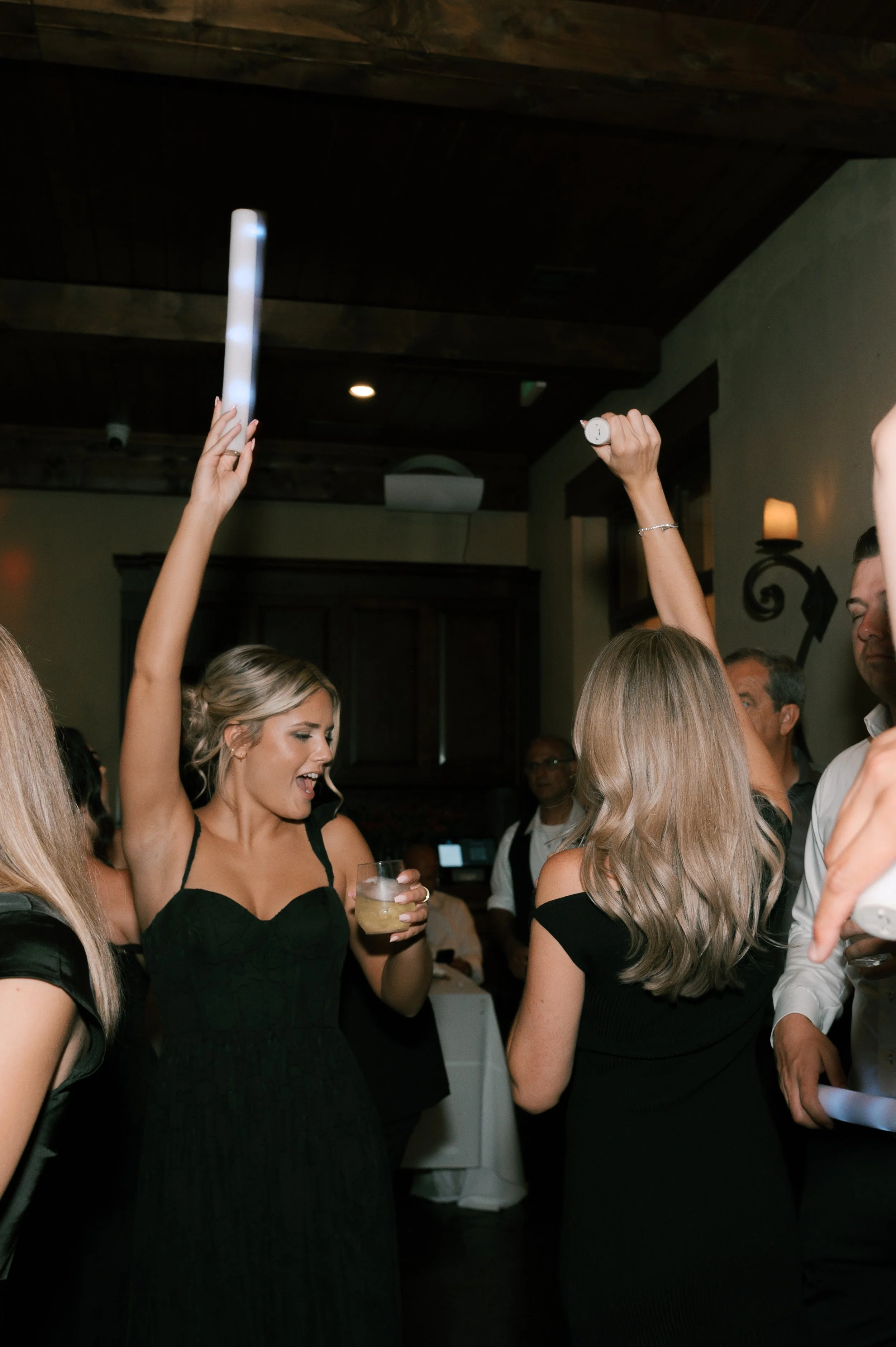 People dancing and celebrating at a party, raising their hands with glow sticks while holding drinks.