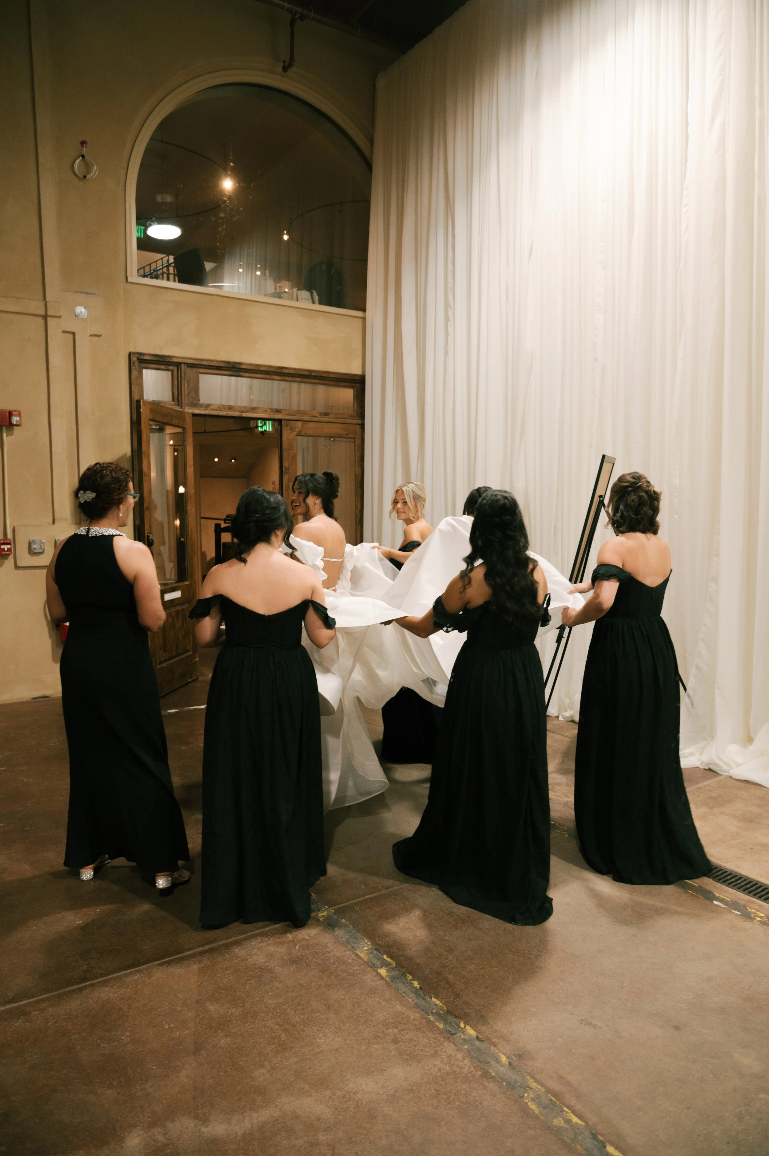 A bride is being helped with her wedding gown by five women in black dresses, in an indoor venue with large white curtains and a high ceiling.