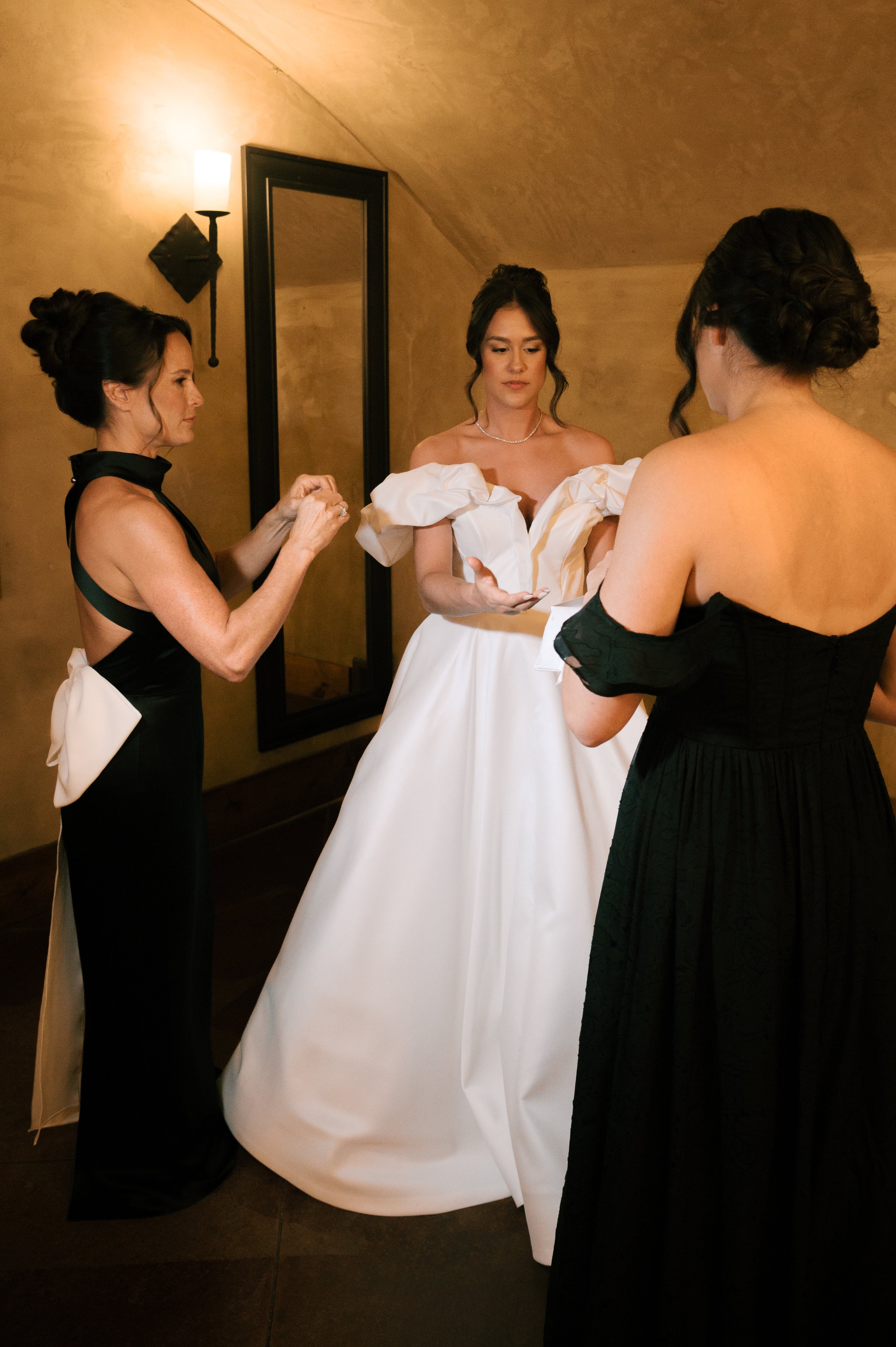 Three women get ready for a wedding, with one wearing a white wedding gown and two in black dresses, in a warmly lit room.