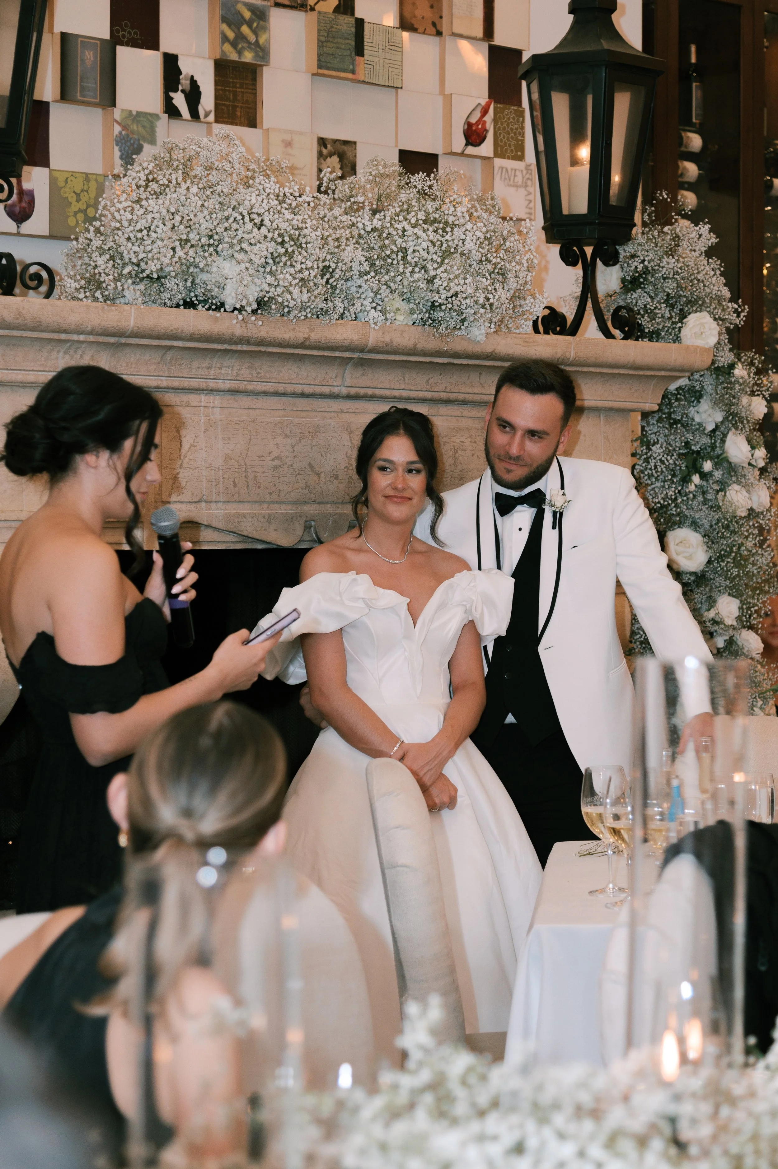 Bride and groom standing at wedding reception, with a woman reading vows or speech, all in formal attire, in a decorated venue with floral arrangements and large fireplace.