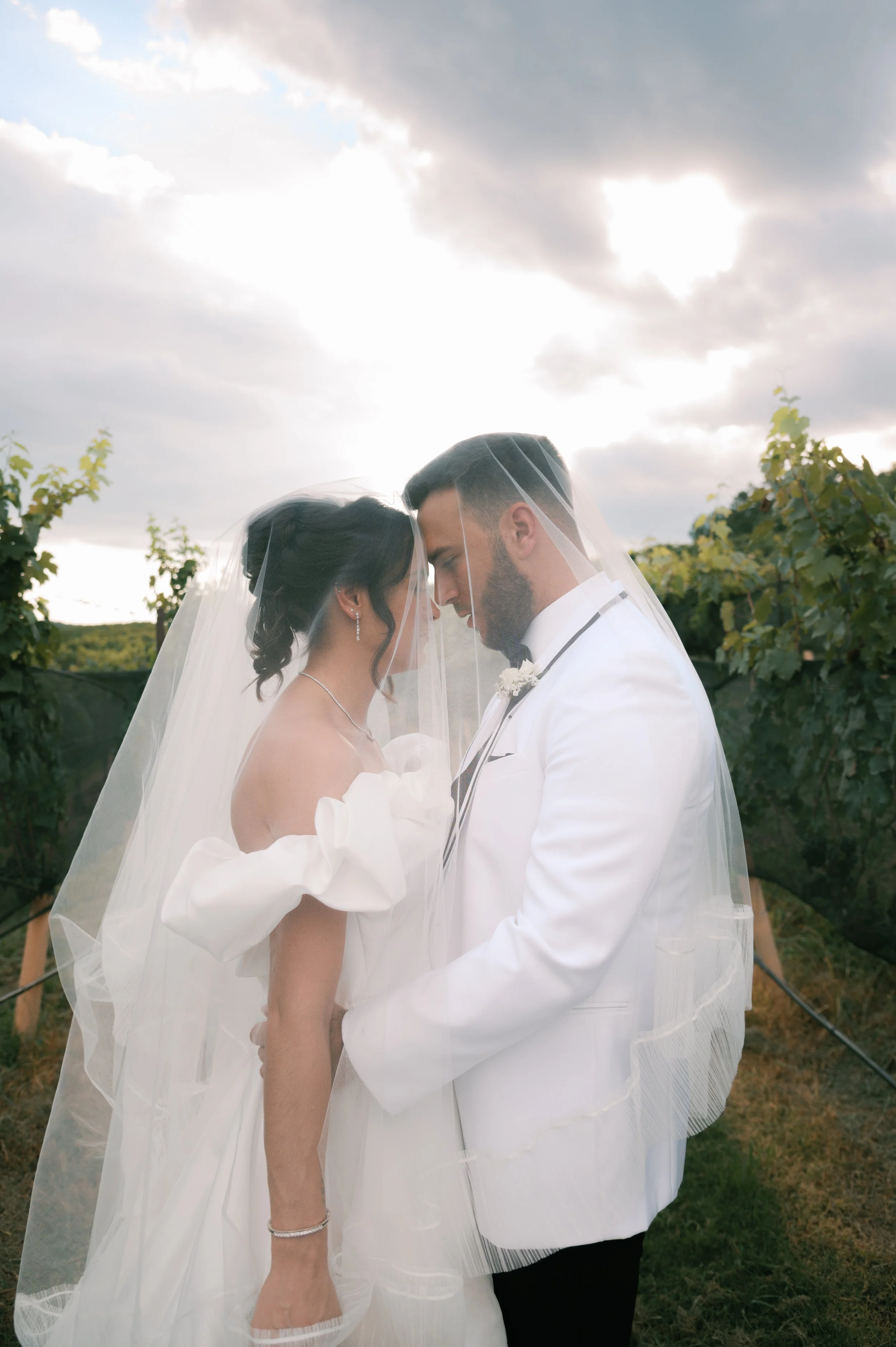 Bride and groom with veils touching faces in a vineyard during sunset.
