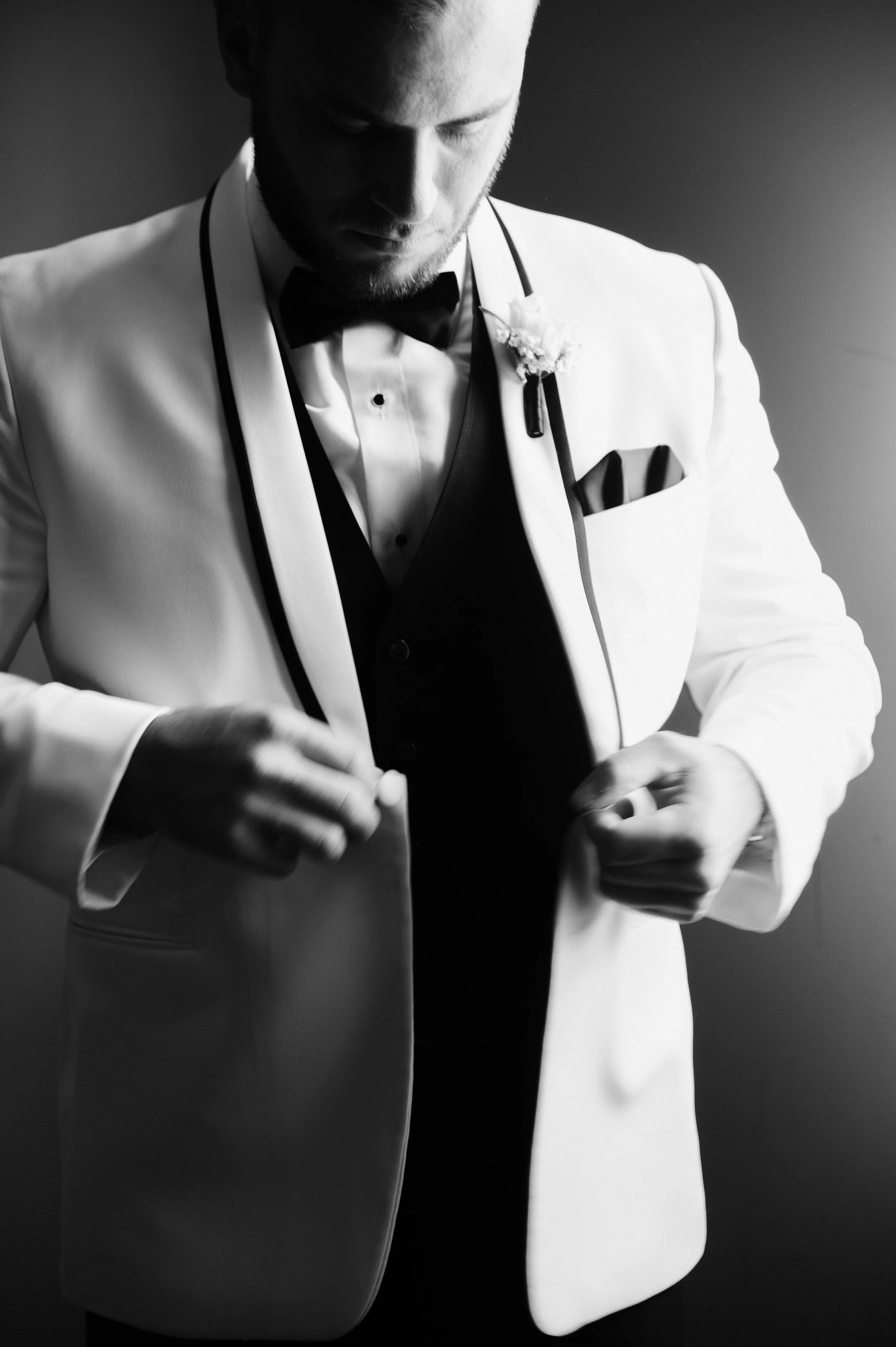 A man in a tuxedo, adjusting his white jacket, preparing for a formal event in black and white photograph.