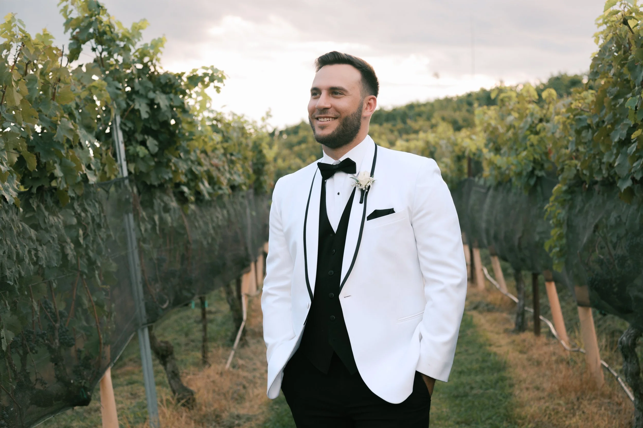Man in a white tuxedo jacket with black trim, black bow tie, and boutonniere standing in a vineyard.