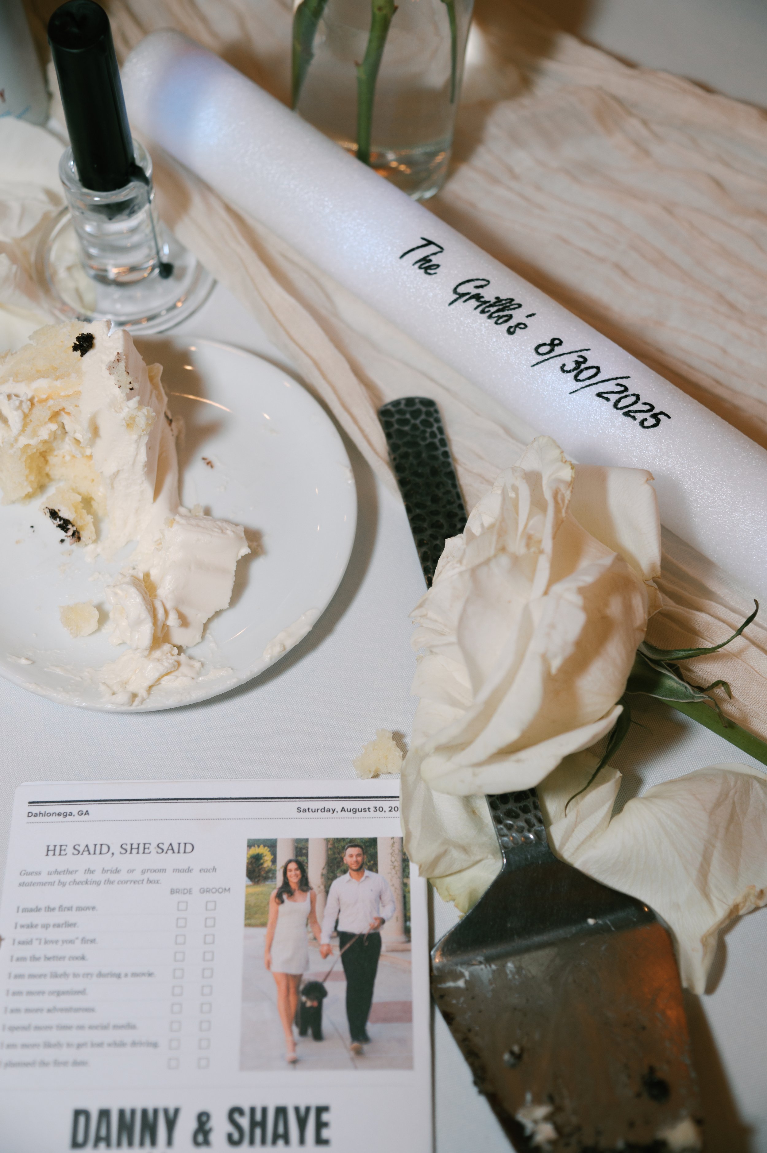 Wedding reception table with a cake partially eaten, a white rose, a sparker, a photo booth picture of a couple with a dog, and a scroll with a date.