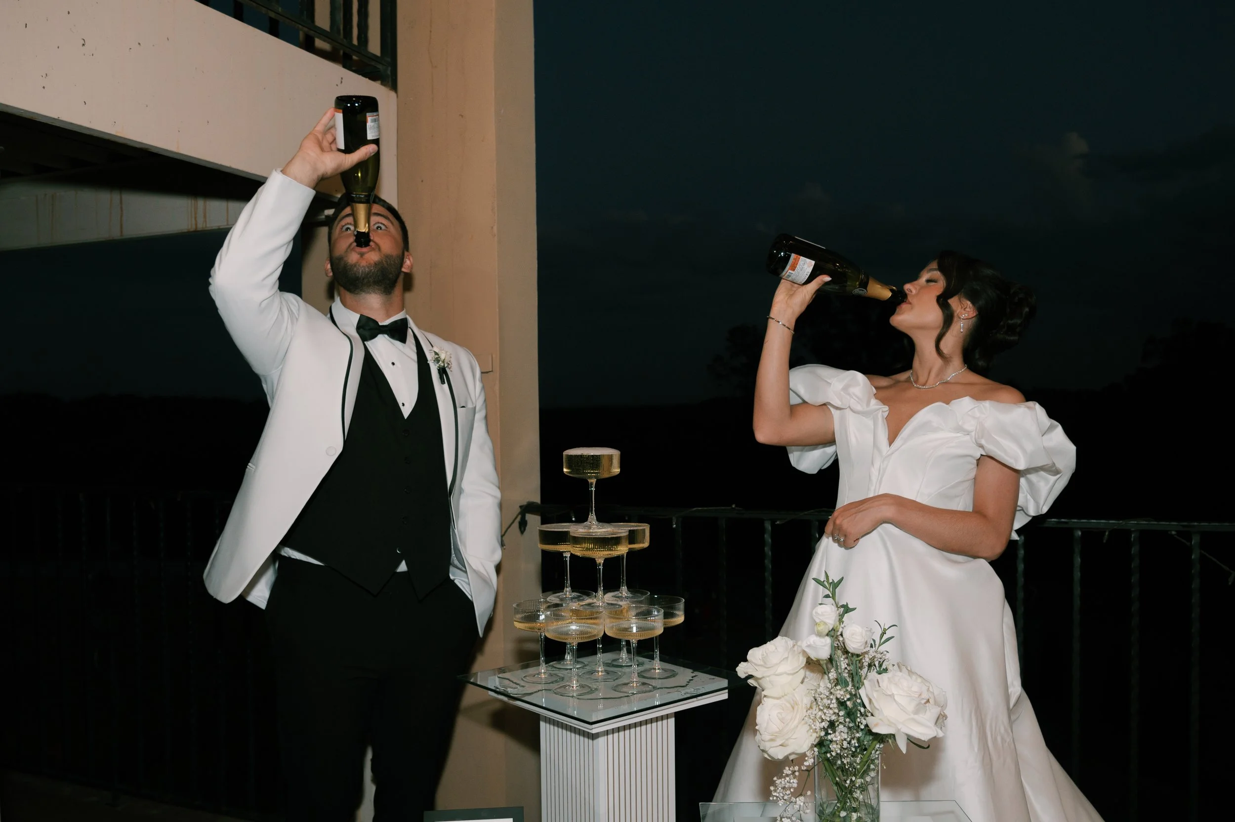 Bride and groom drinking champagne at a wedding reception, standing outdoors during nighttime. The groom in a white tuxedo jacket and black bowtie, and the bride in a white wedding dress with puff sleeves, holding bouquets of white roses.