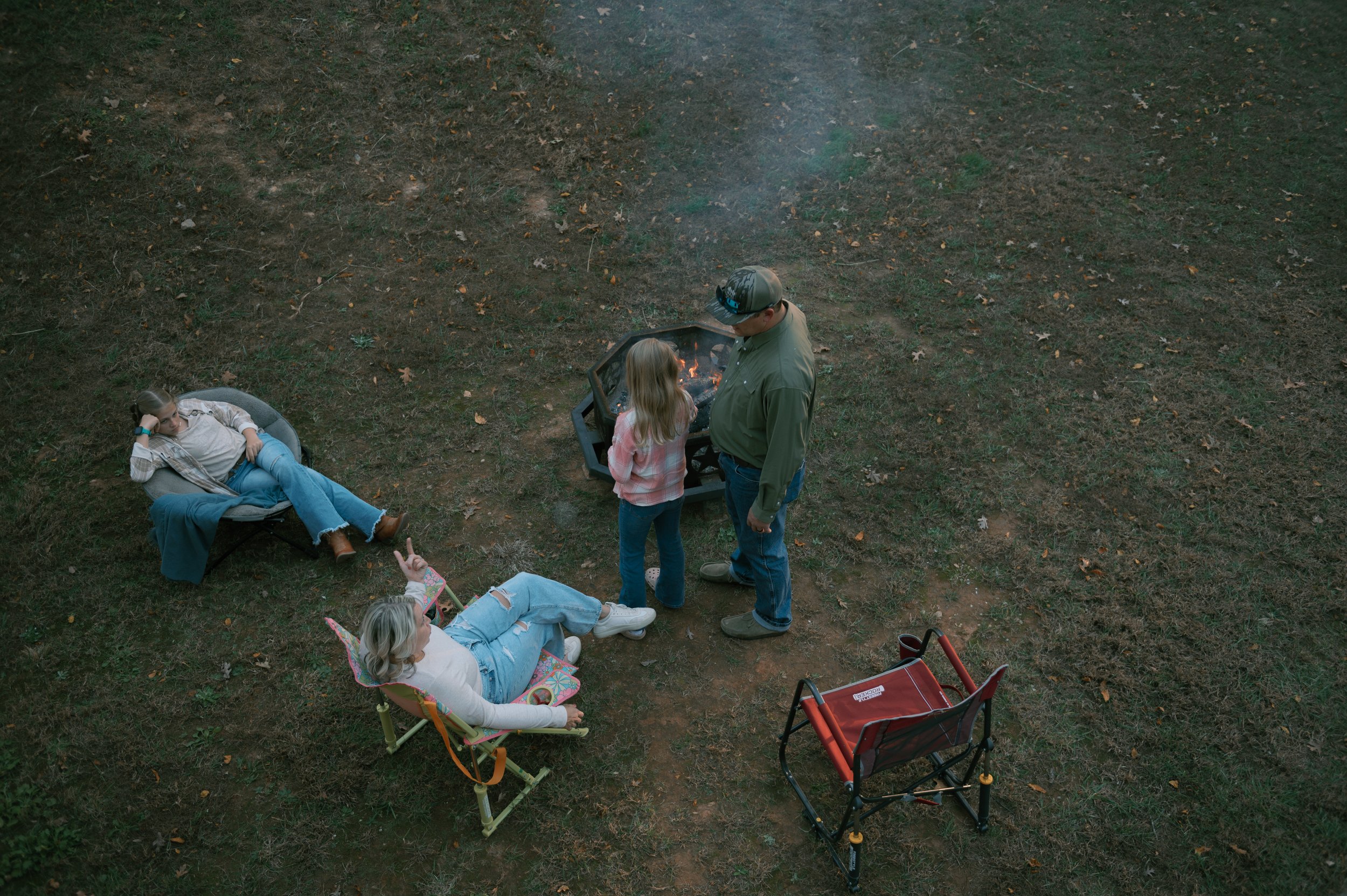 A group of people gathered around a small fire pit in a wooded area, some sitting on portable chairs and others standing, during what appears to be the evening or late afternoon.