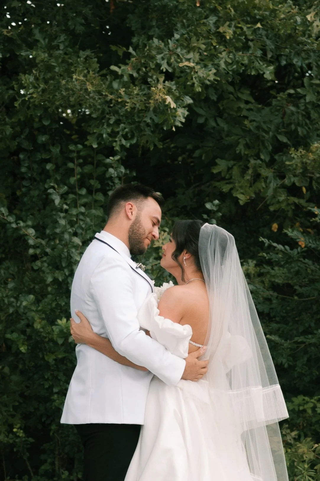 A bride and groom embracing outdoors, standing close together with their foreheads touching, surrounded by green foliage.