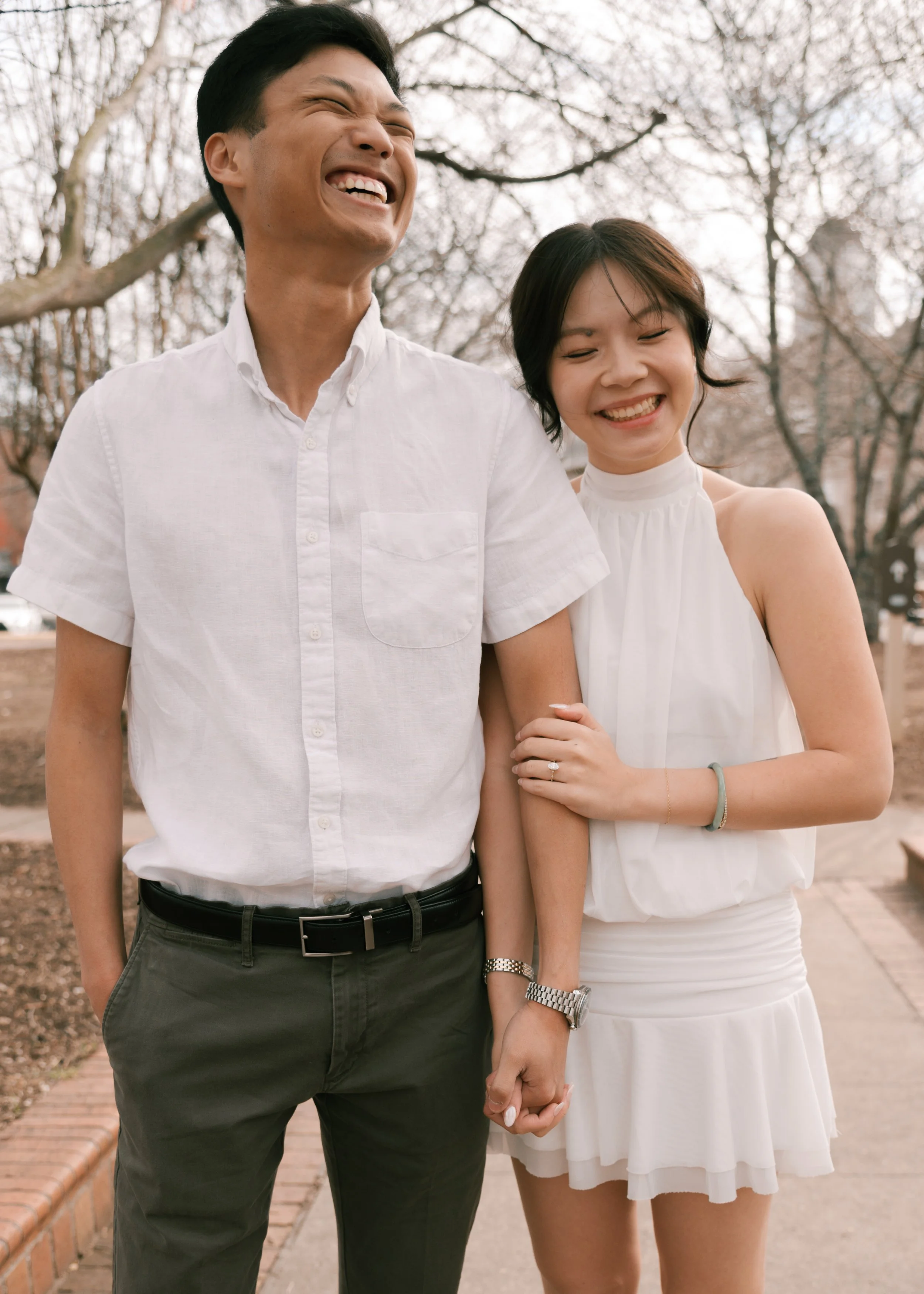 A happy couple walking outdoors on a sunny day, holding hands and smiling at each other, surrounded by trees.