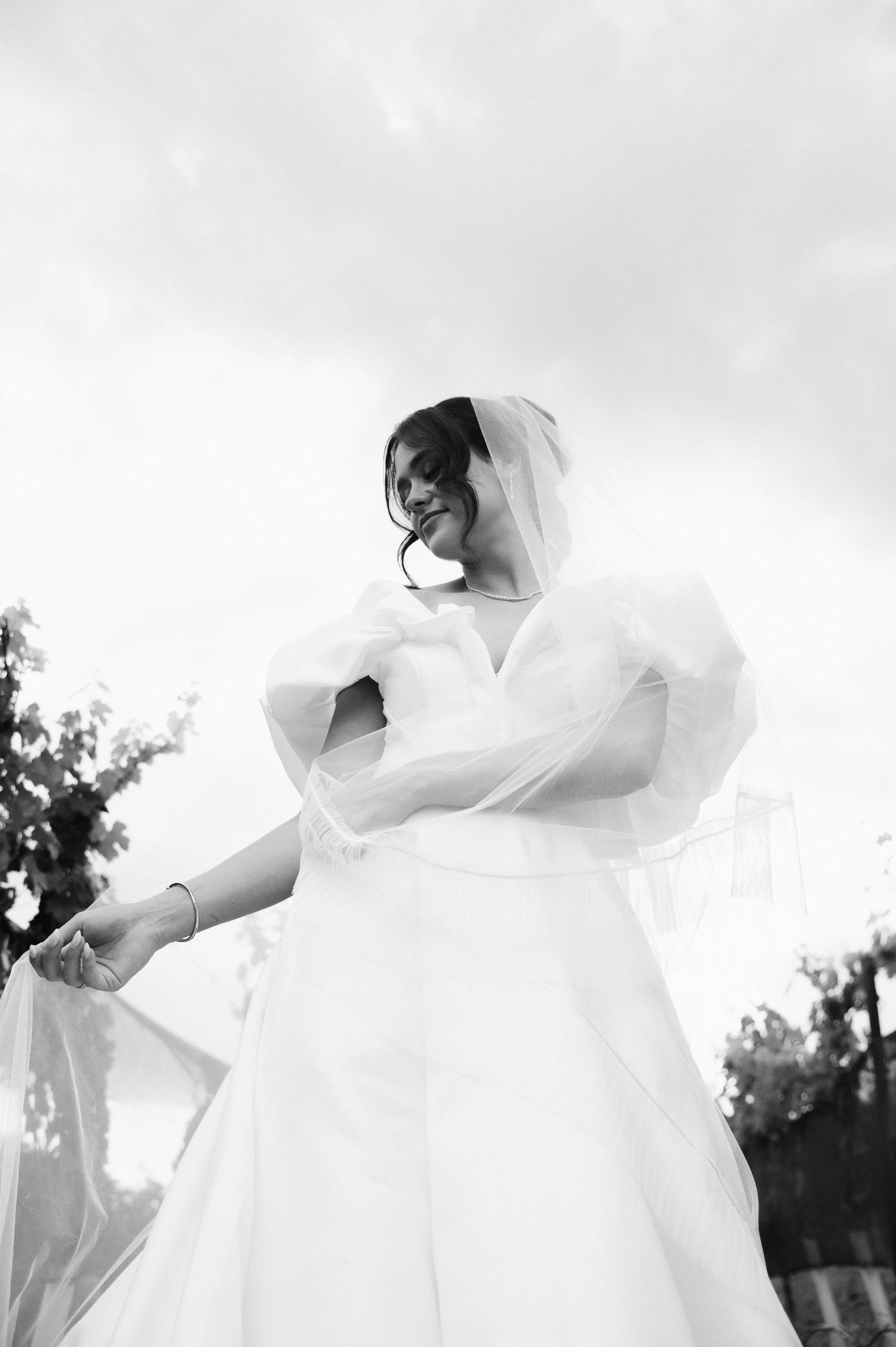 A woman in a wedding dress looking down, taken from a low angle in black and white, outdoors.