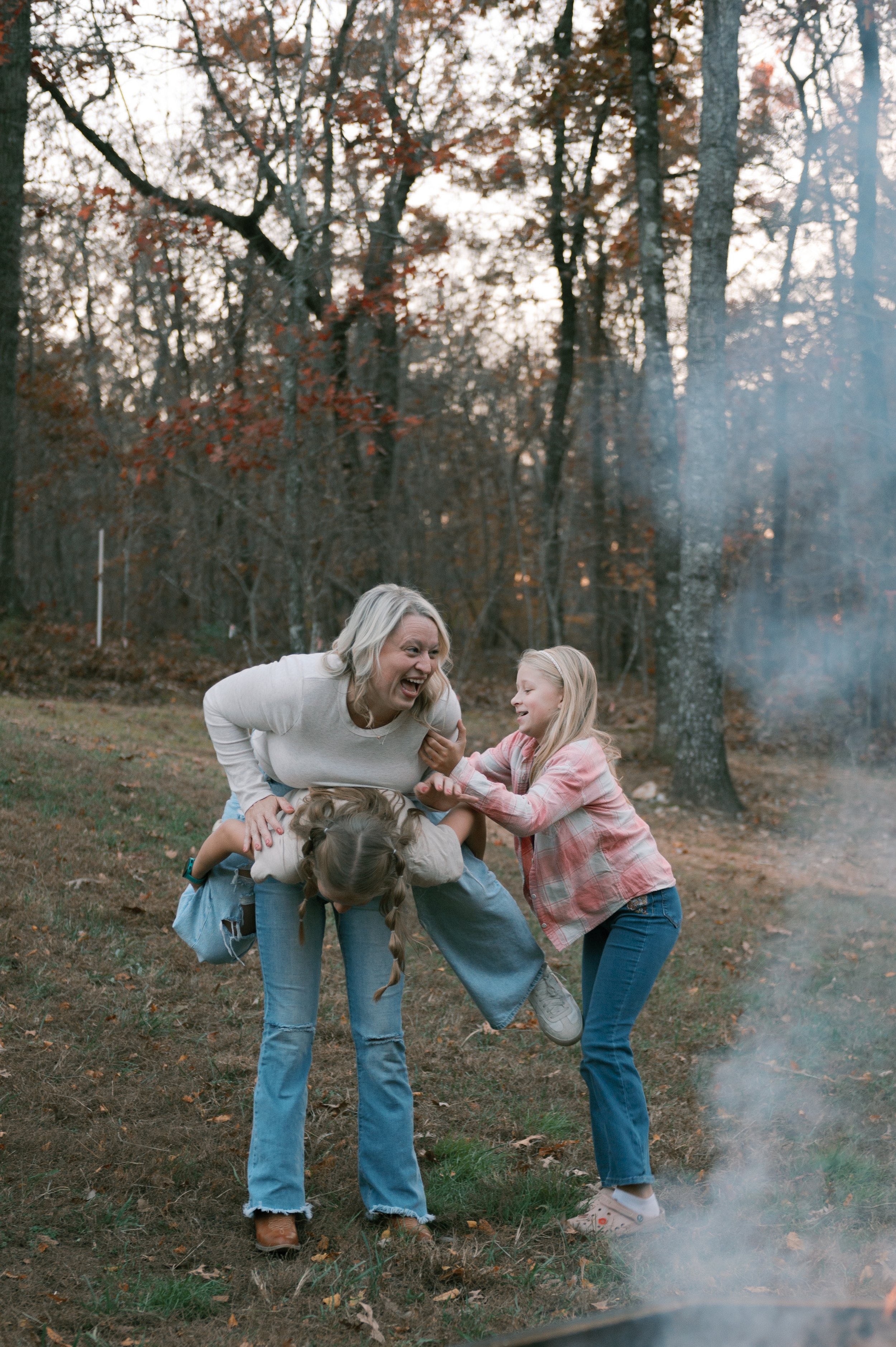 A group of people, including a woman and children, outdoors in a wooded area, playing and laughing; leaves and trees in the background, with a light smoke or mist nearby.