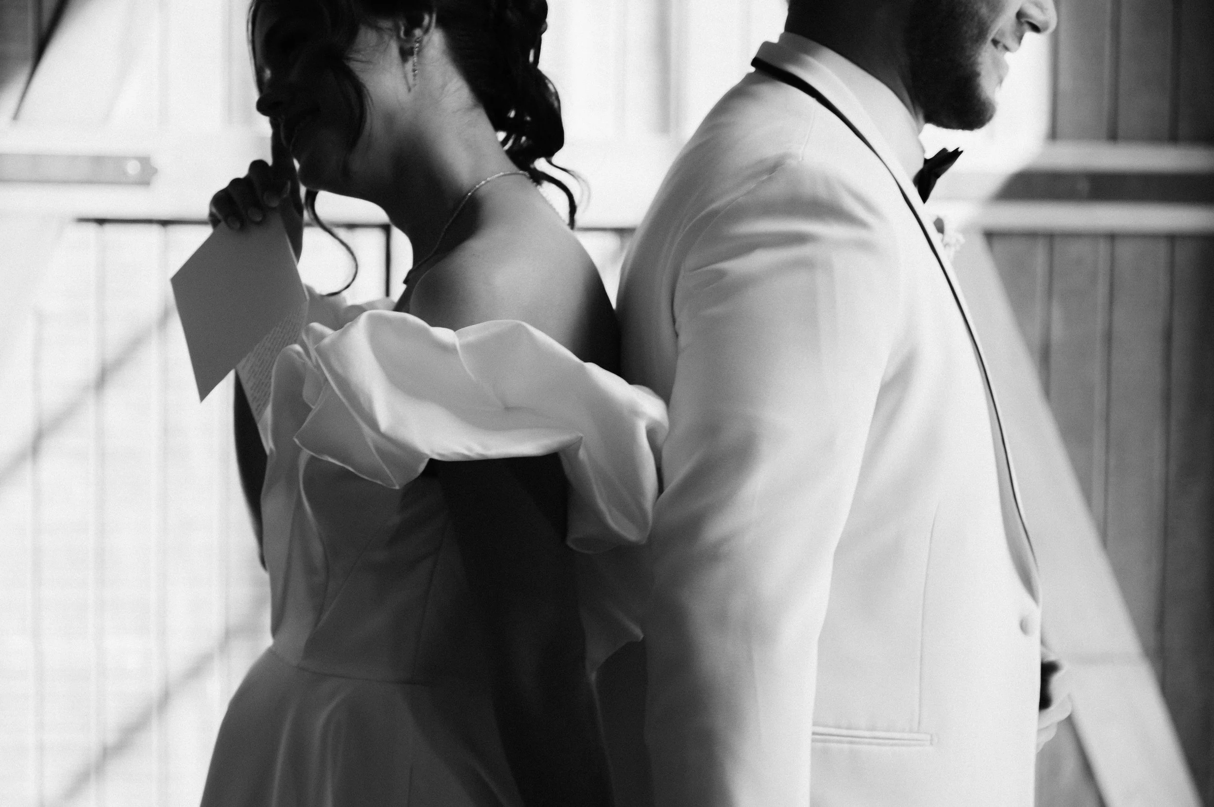Black and white photo of a couple in wedding attire standing back-to-back in a barn-like setting.