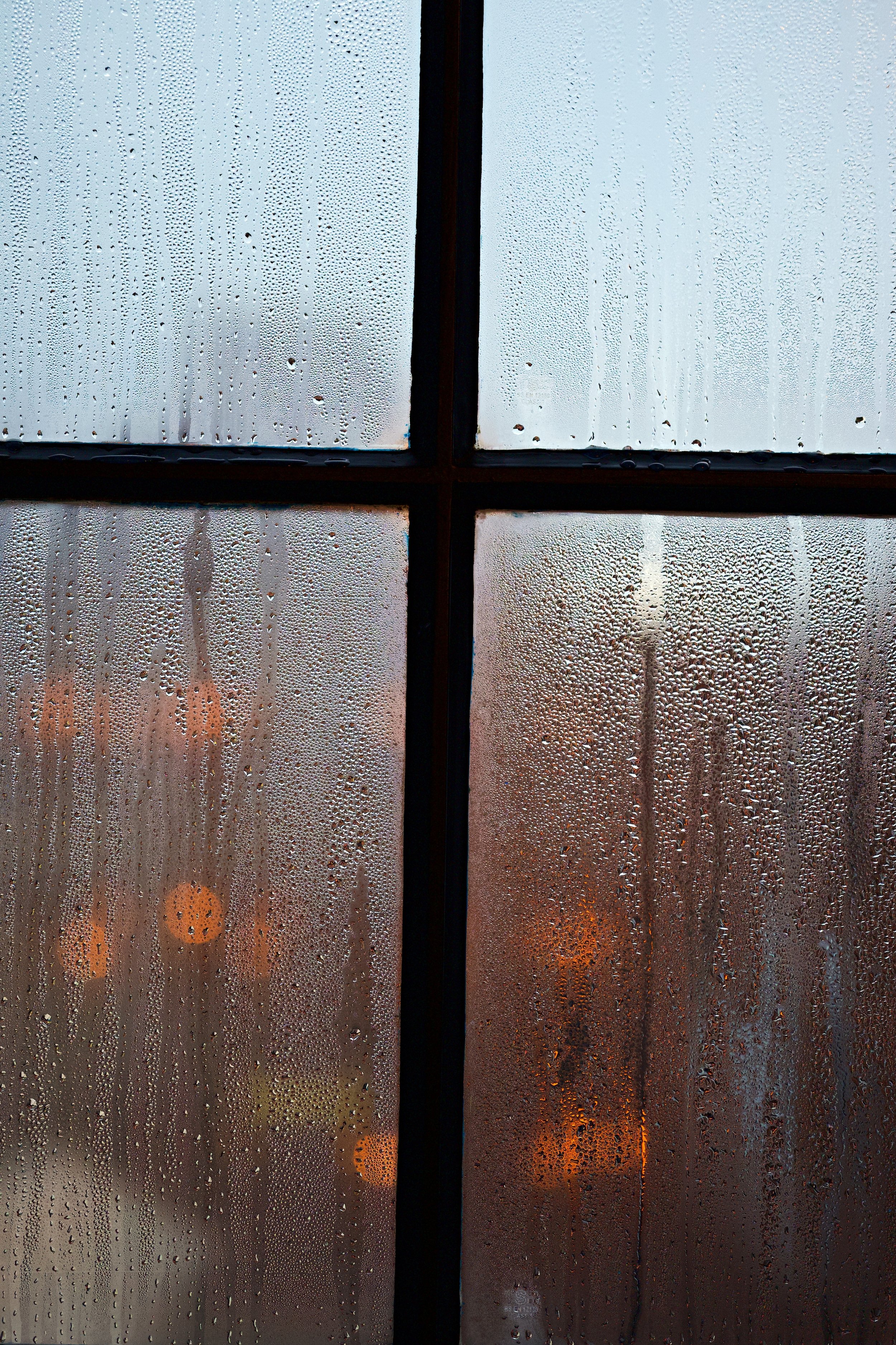 Rain-covered window with droplets on glass, showing blurred city lights and buildings outside at dusk or dawn.