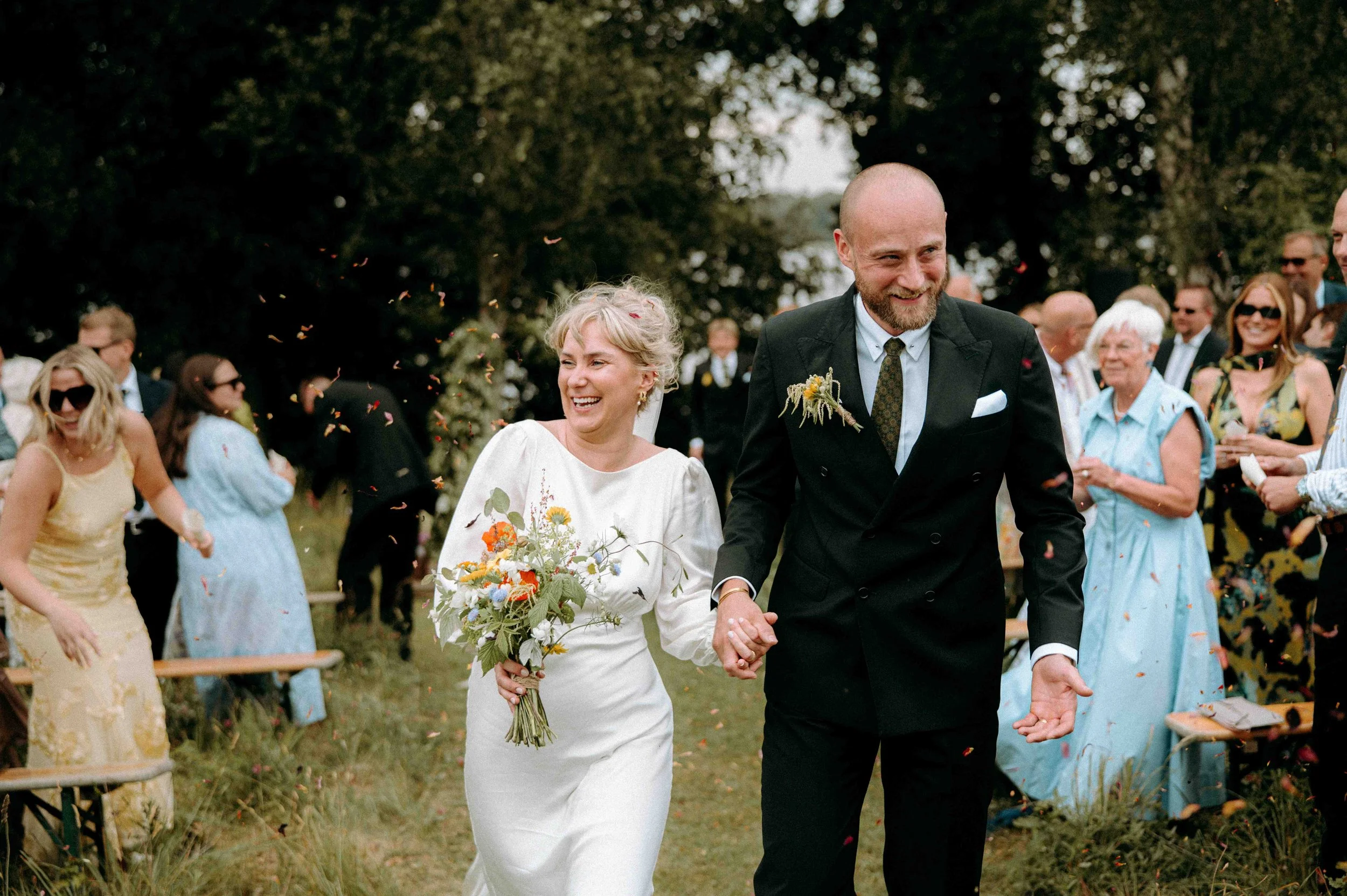 Två röda trähus med tegel- och mossbeklädda tak, ett med liten veranda och ett annat med ett rödmålat fönster, omgivna av träd och växtlighet i höstfärger.