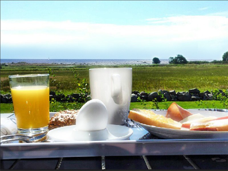 Frukost på ett bord med ägg, juice, kaffe och bröd, utsikt över grönt landskap och hav i bakgrunden.