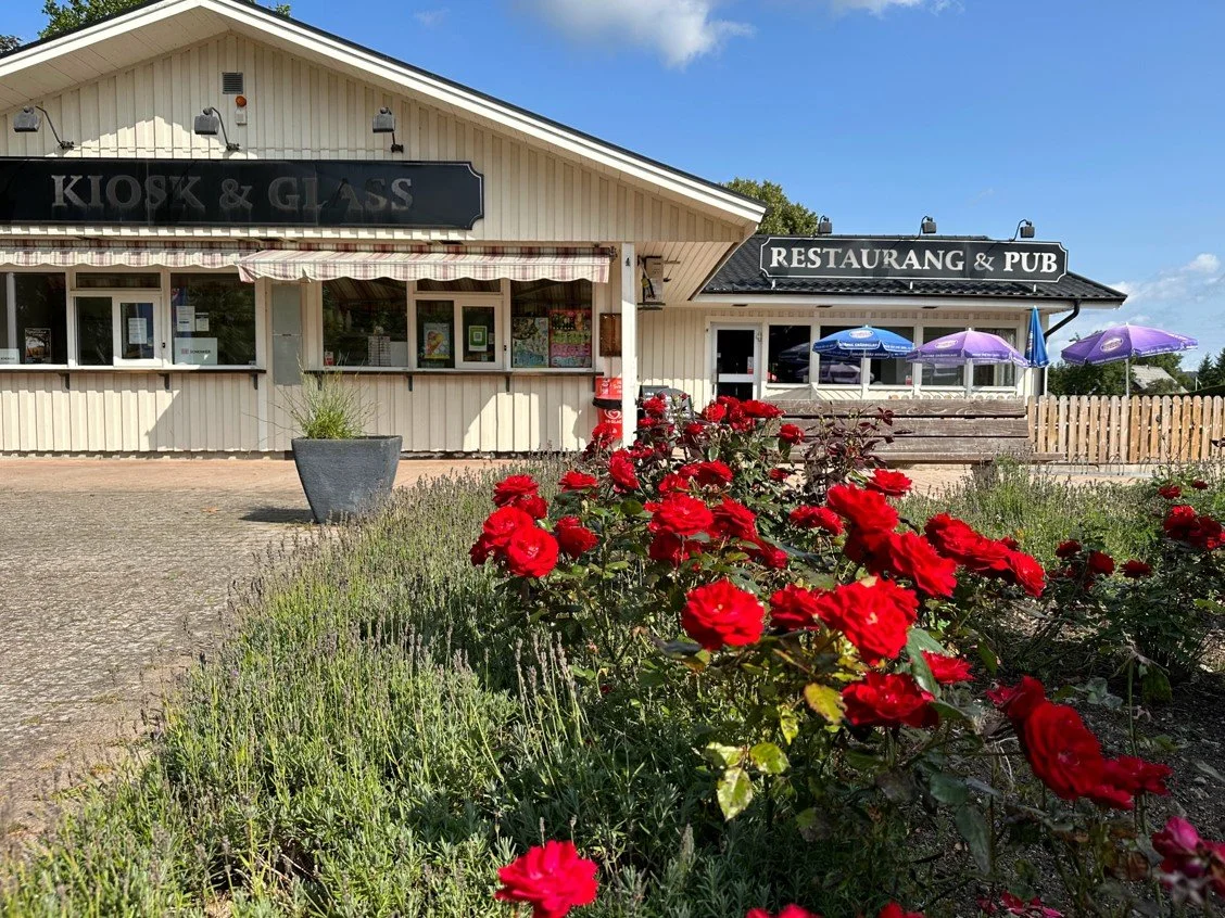 Framsidan av en restaurang och pub med blomsterbädd med röda blommor i förgrunden. En byggnad med vita väggar och skyltar som säger 'Kiosk & Glass' samt 'Restaurang & Pub'. Under ett av takskydden finns flera parasoller i lila och blå.