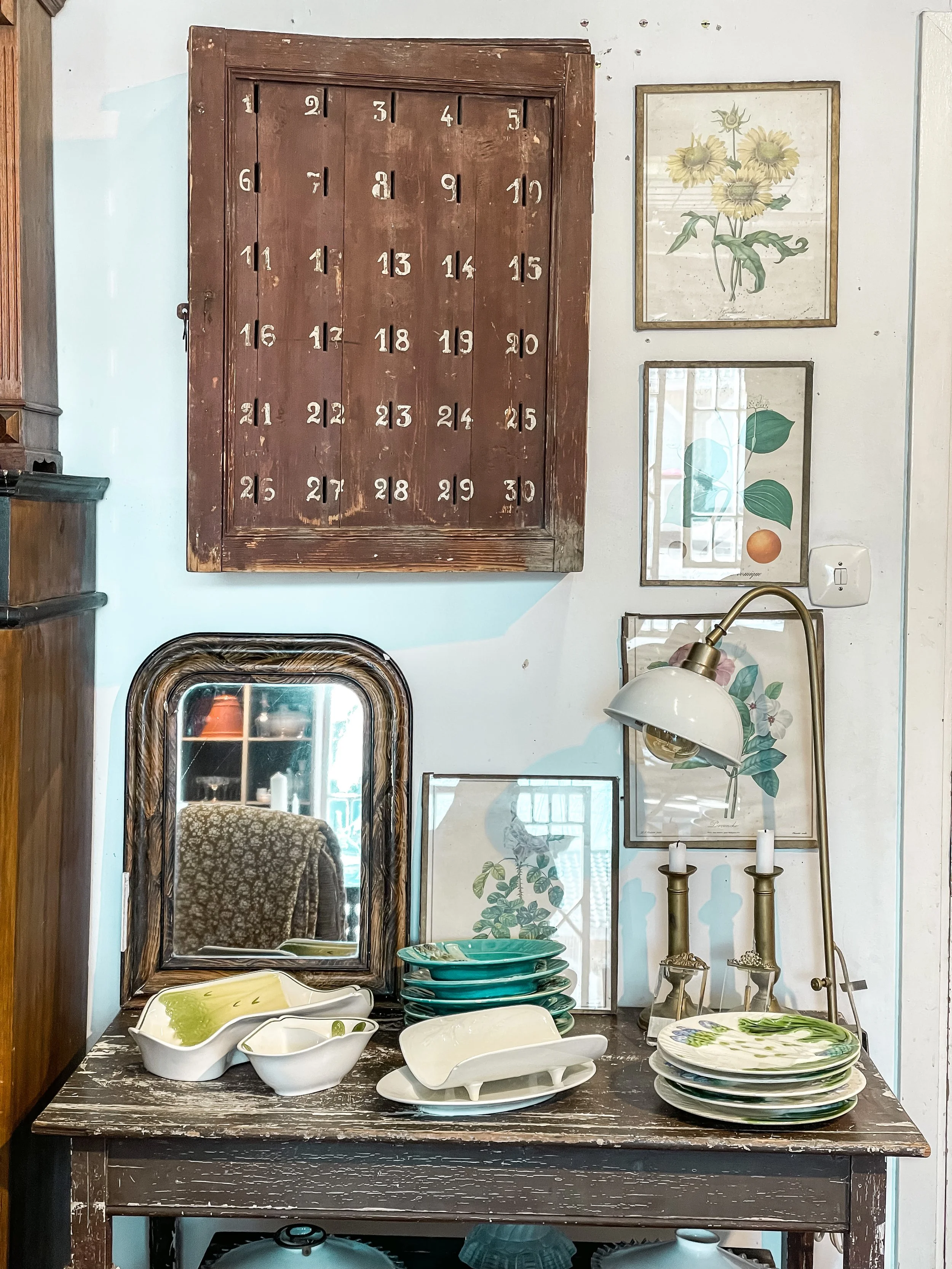 En träbord med gröna tallrikar och vita skålar, spegel och bilder på växter på väggen, samt en väggalmanacka.