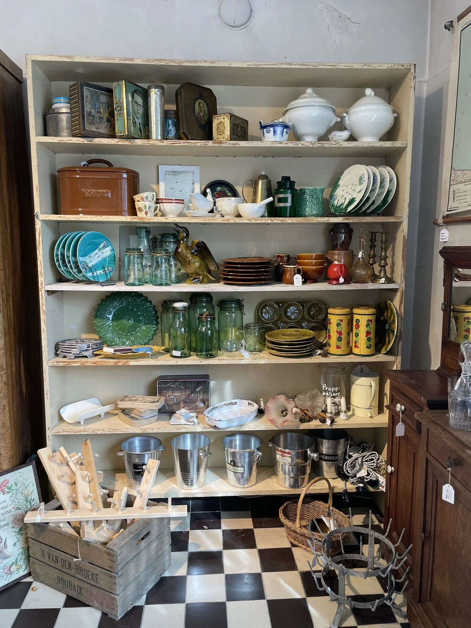 Vitt köksskåp fyllt med vintage porslin, glas, burkar och ett antal andra köksartiklar, med svart och vitt rutigt golv.