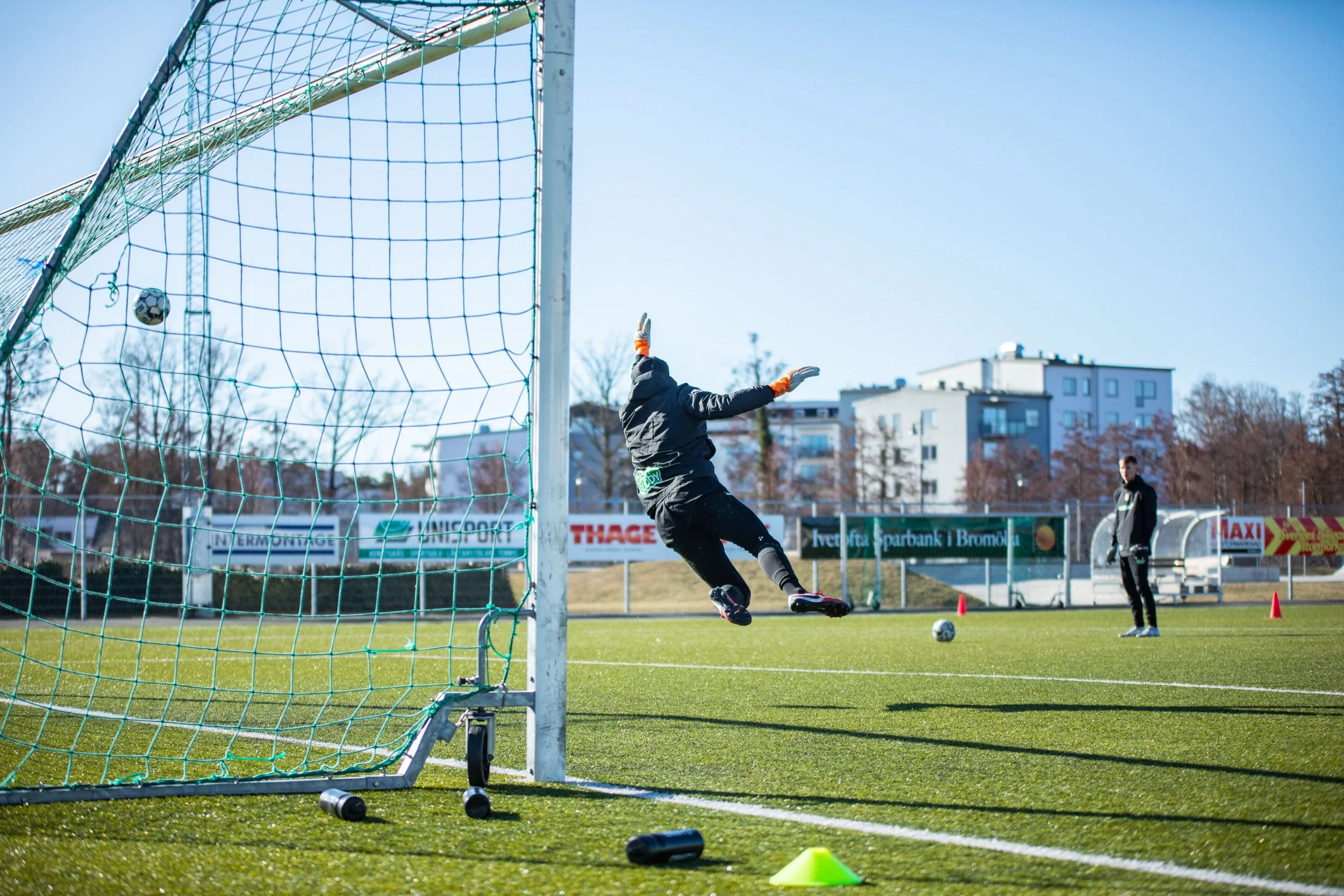 Fotbollstränare tränar en målvakt på en fotbollsplan en solig dag.
