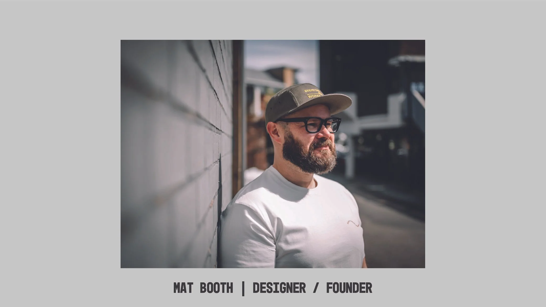 Man with glasses and a beard wearing a baseball cap and white t-shirt, standing outdoors leaning against a tiled wall.