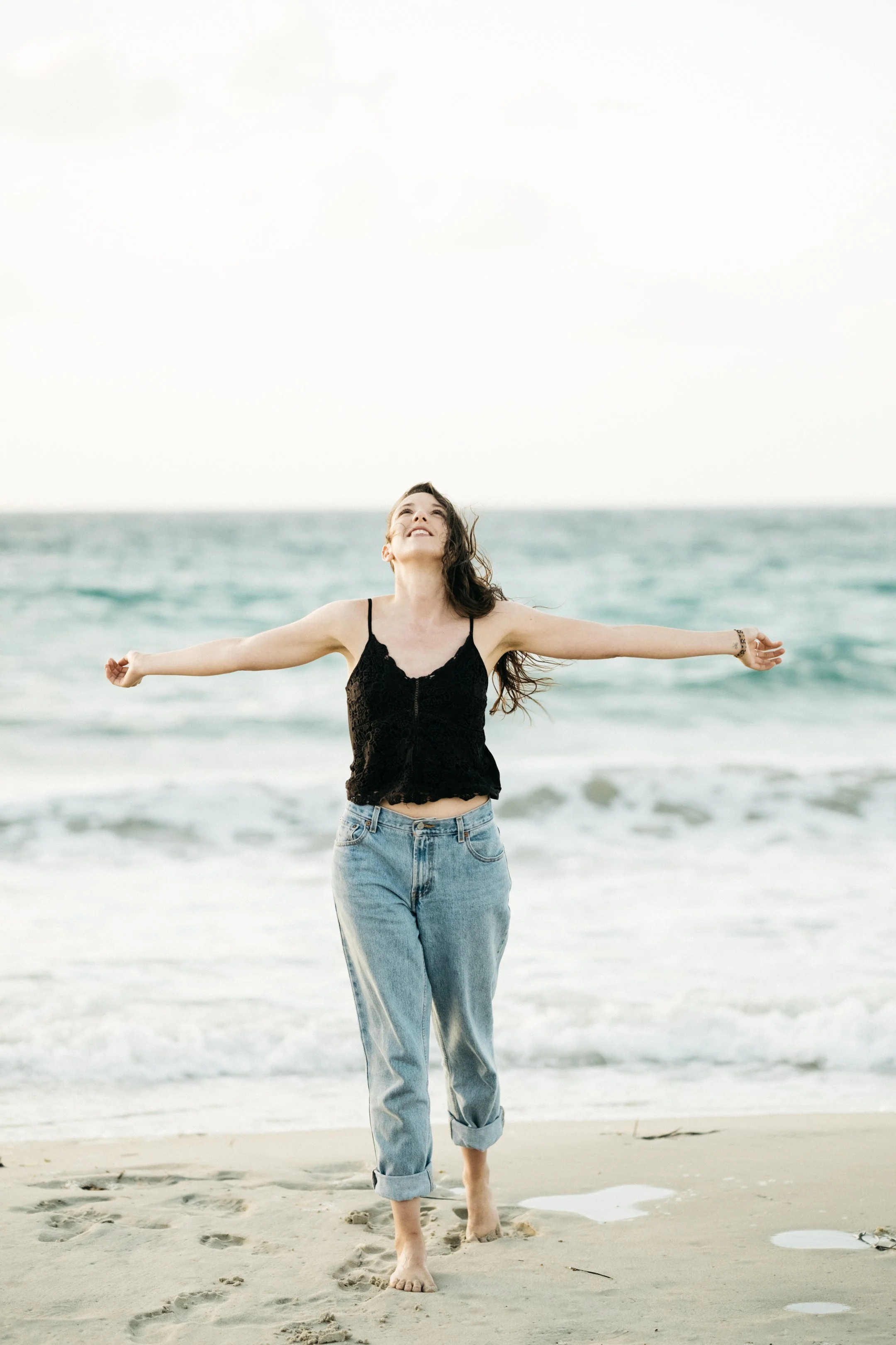 Woman with long hair wearing black top and rolled-up jeans walking barefoot on the beach near the ocean, arms outstretched, smiling with sea in background.