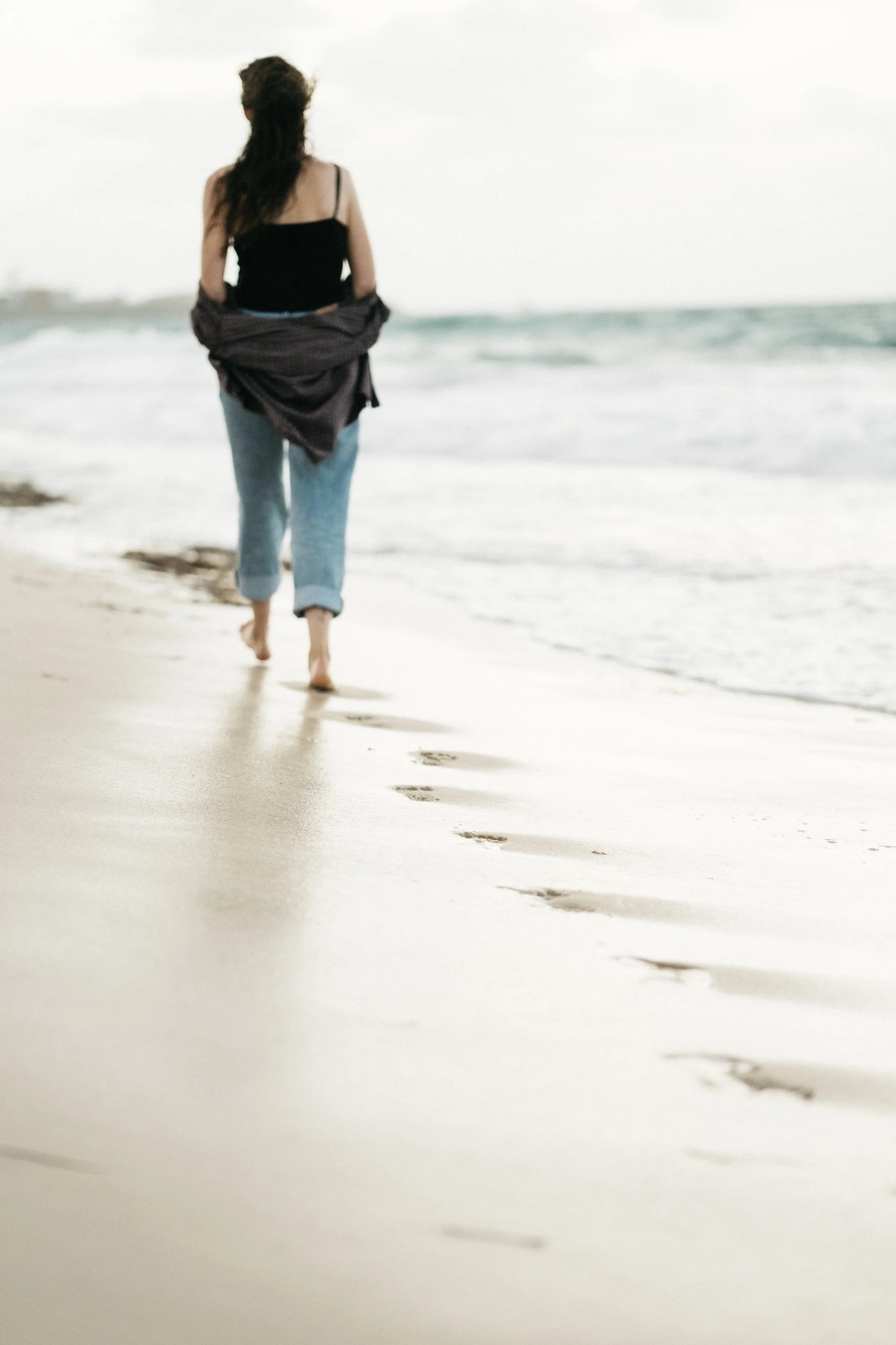 A woman walking along the shoreline at the beach, leaving footprints in the sand, with the ocean waves in the background during a cloudy day.