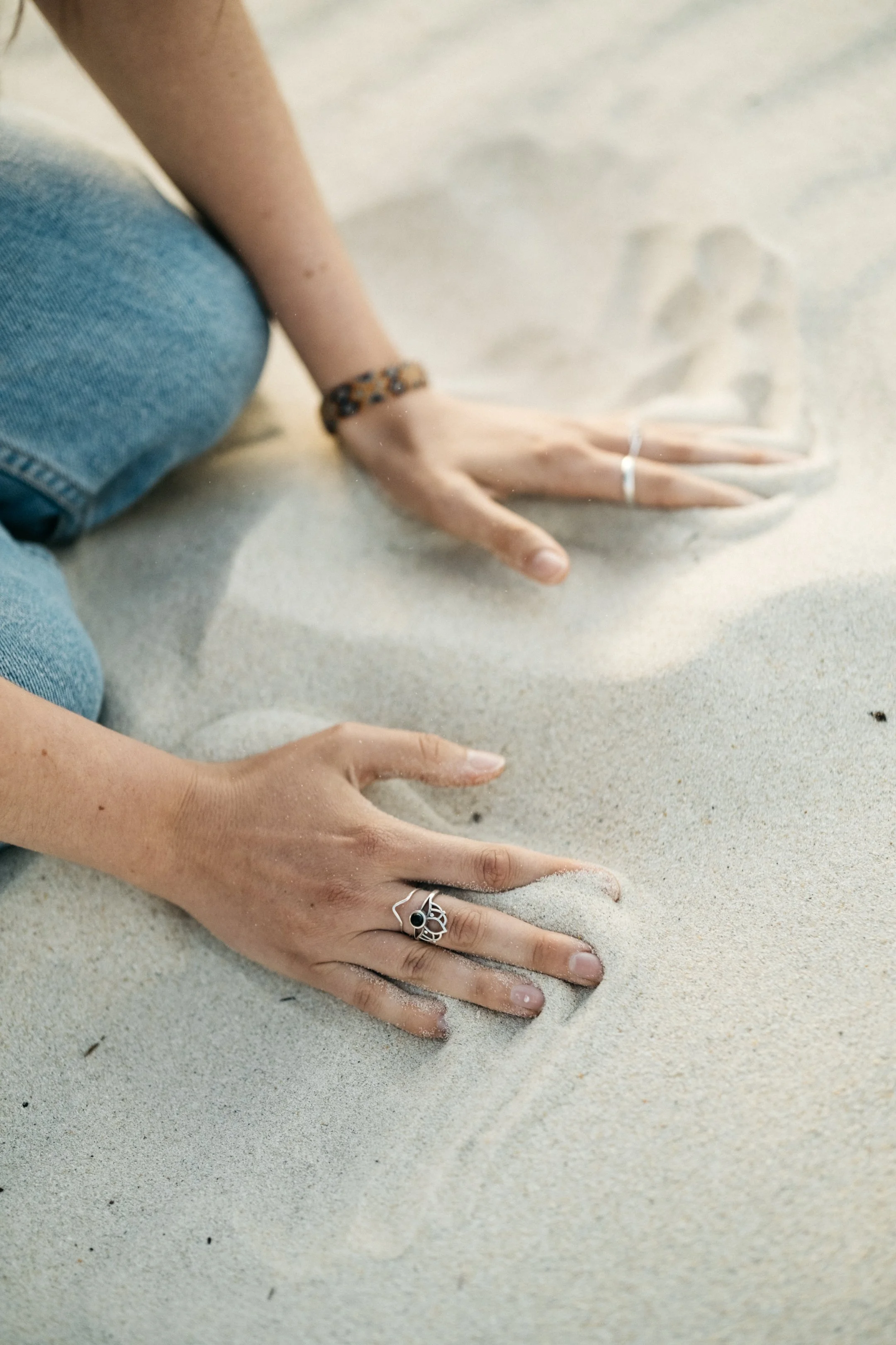Person's hands and knees on sandy beach, wearing rings and bracelets, with sand between fingers.