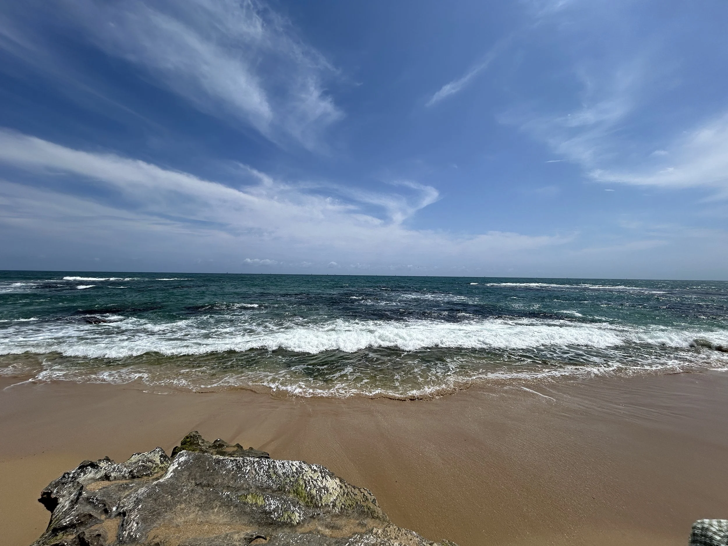 View of the ocean with waves crashing onto a sandy beach and a partly cloudy blue sky.
