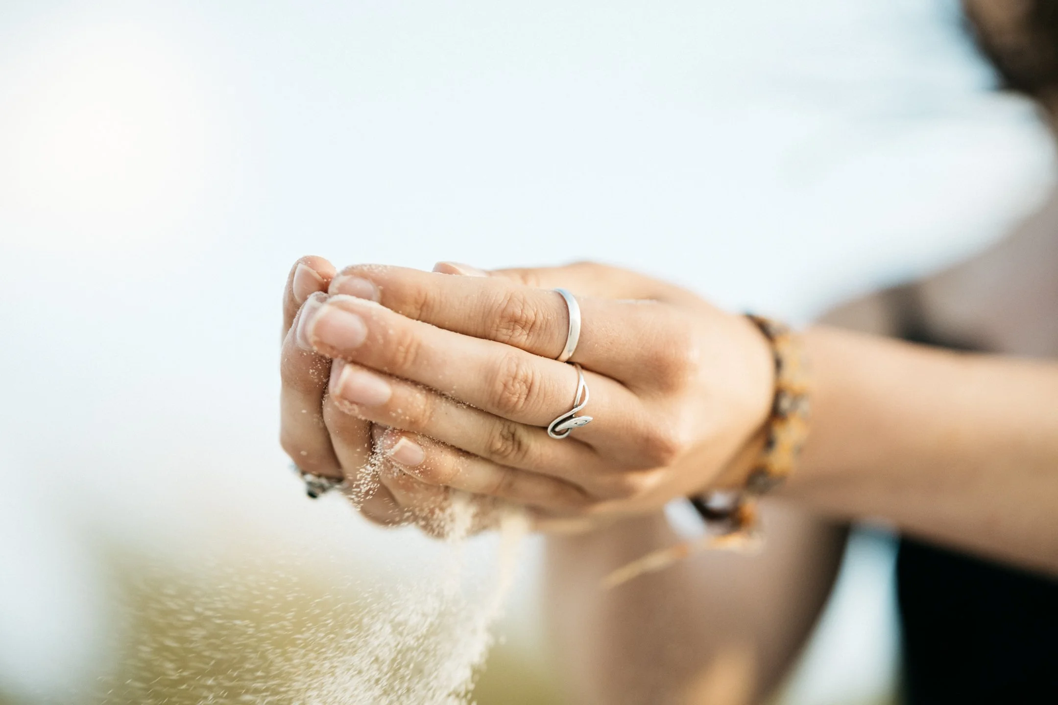 Person's hands pouring white sand through fingers, wearing rings and bracelets, with a blurred outdoor background.