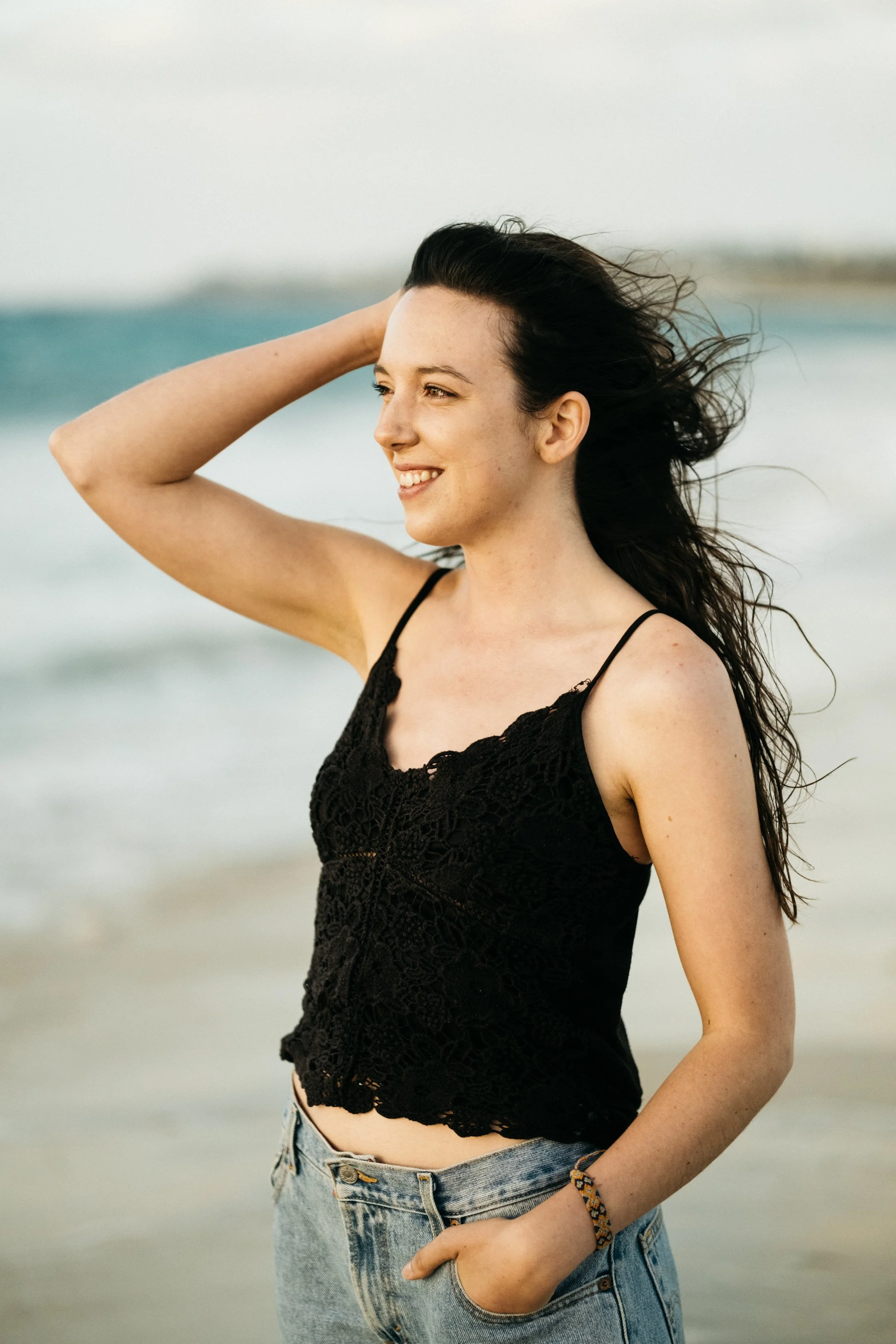 A young woman with long dark hair smiling at the beach during sunset, wearing a black lace top and jeans.
