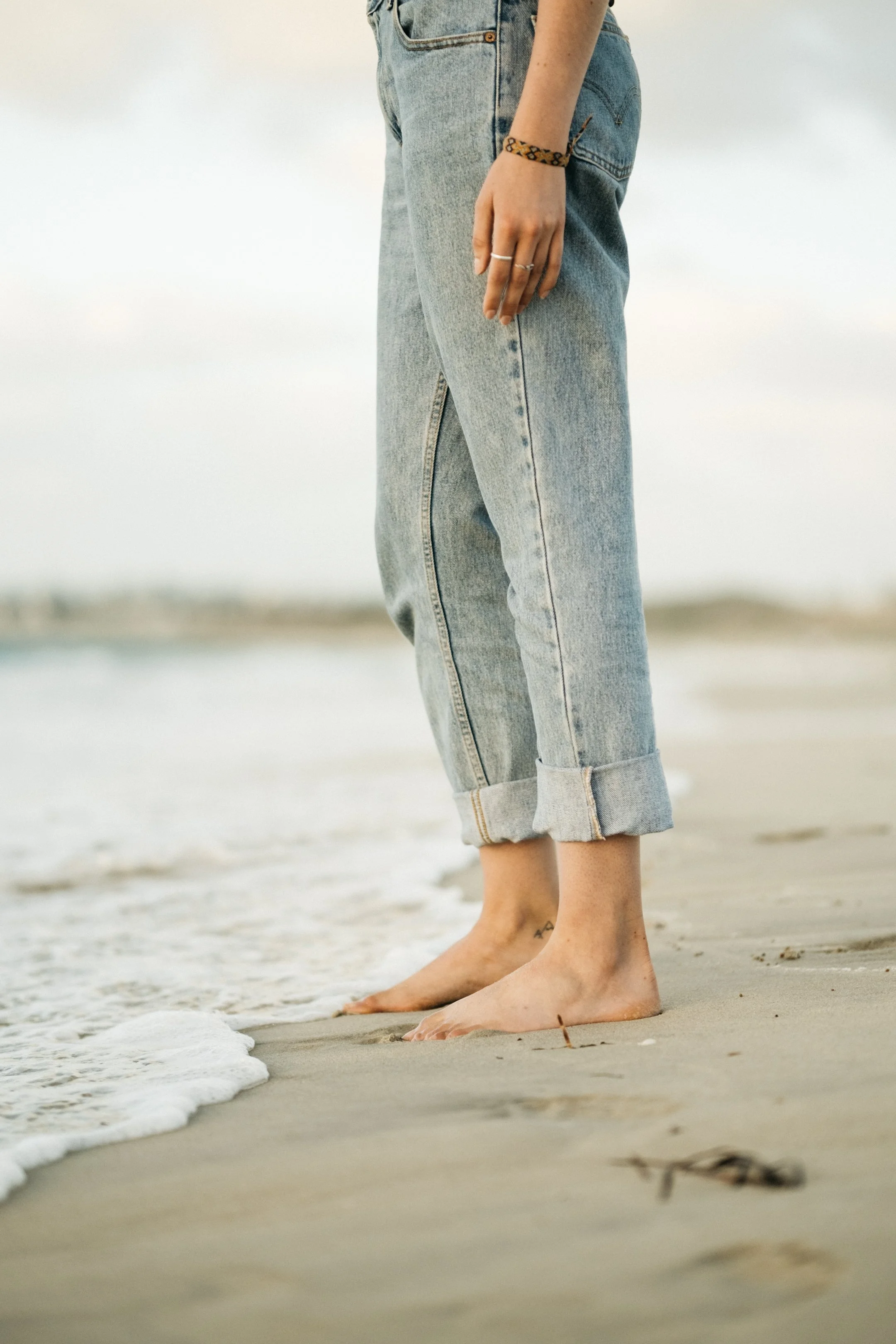 Person standing barefoot on the sand at the beach, wearing rolled-up jeans and a bracelet.