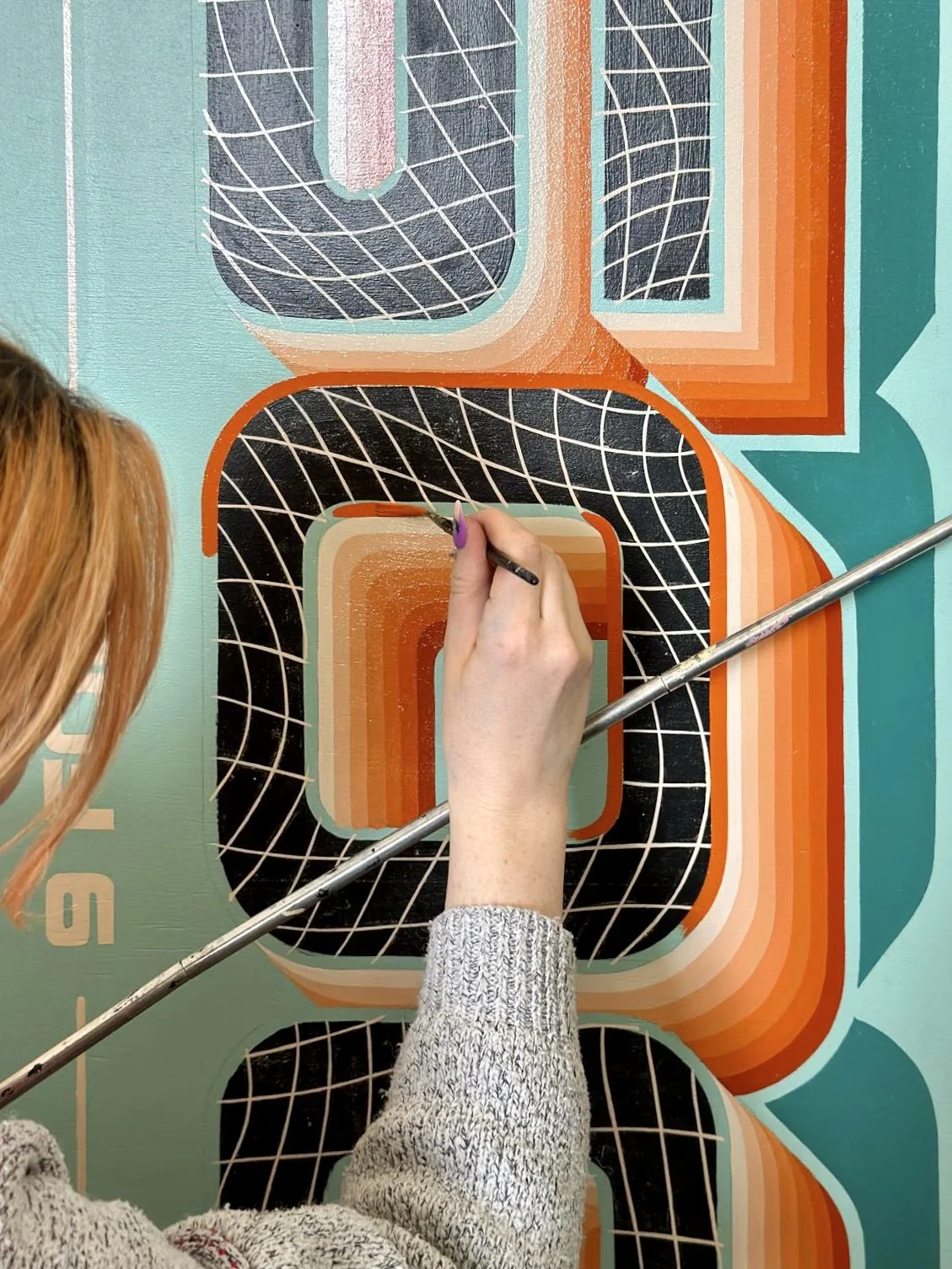 Artist painting a colorful, geometric, complex sign with black, orange, teal, and white patterns.