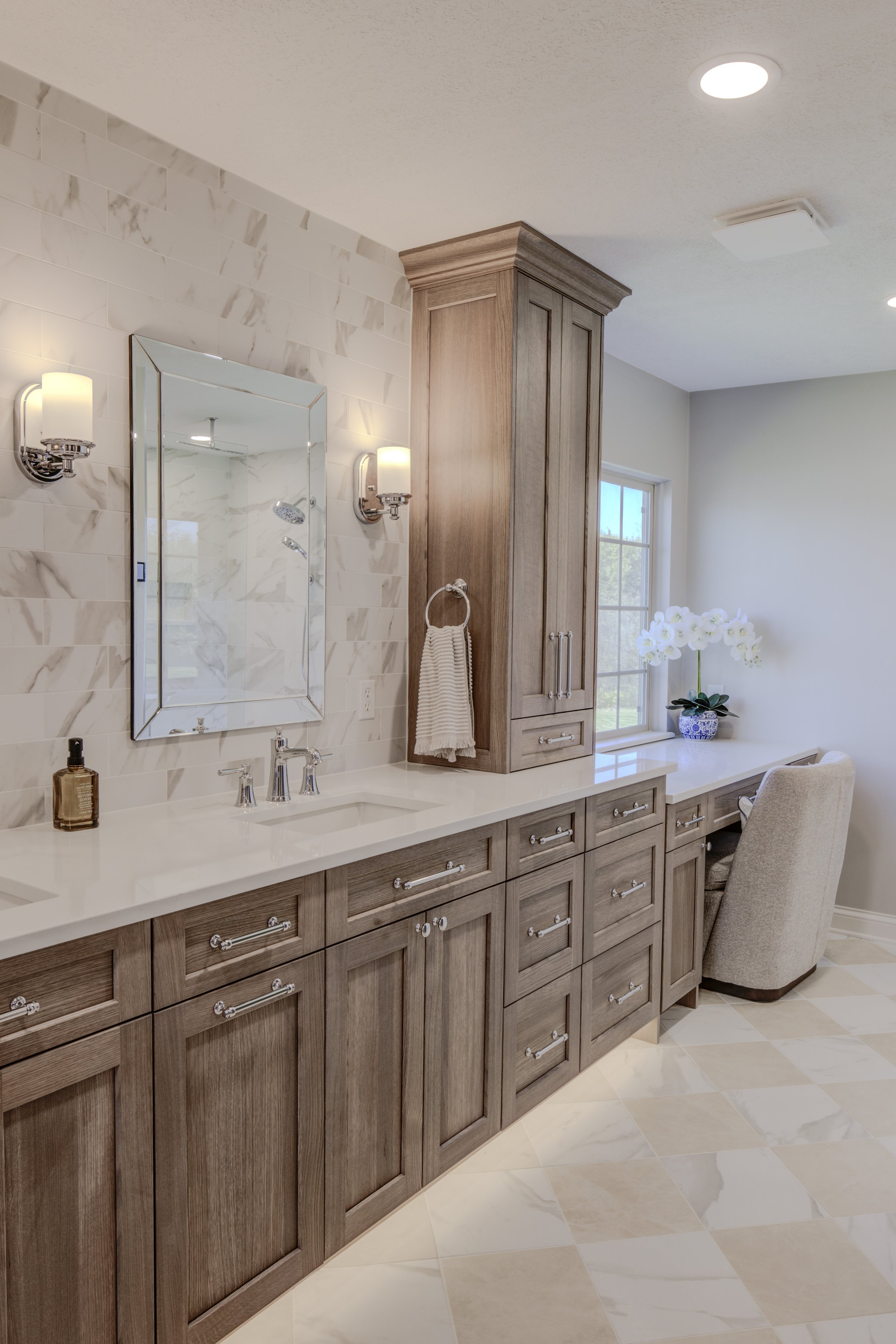 A bathroom vanity with a white countertop, multiple wooden drawers, and cabinets. Behind it, a wall with marble tiles, a mirror, and two wall-mounted lights. To the right, a wooden cabinet, a window with a foliage view, and a vase of white orchids on