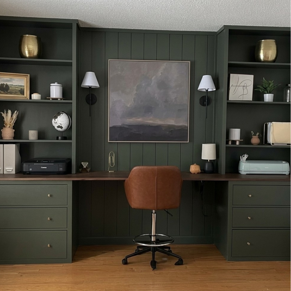 Custom dark green built-in home office unit featuring symmetric tall bookshelves, lower storage drawers, a matching wood-paneled accent wall with mounted sconces, and an integrated double desk workspace.