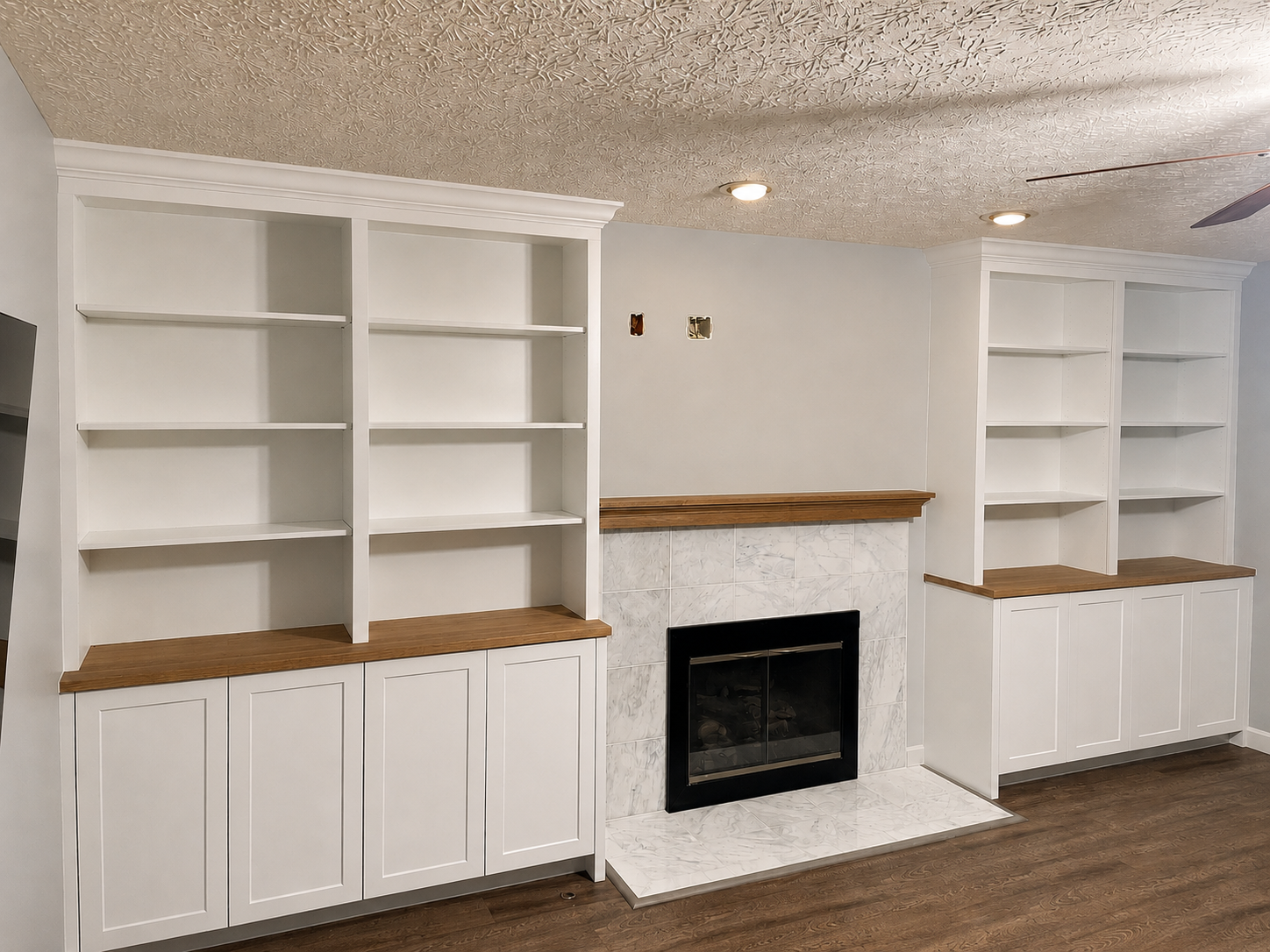 Living room with built-in white bookshelves, a fireplace with a marble surround, and wooden countertops, with a textured ceiling and hardwood flooring.