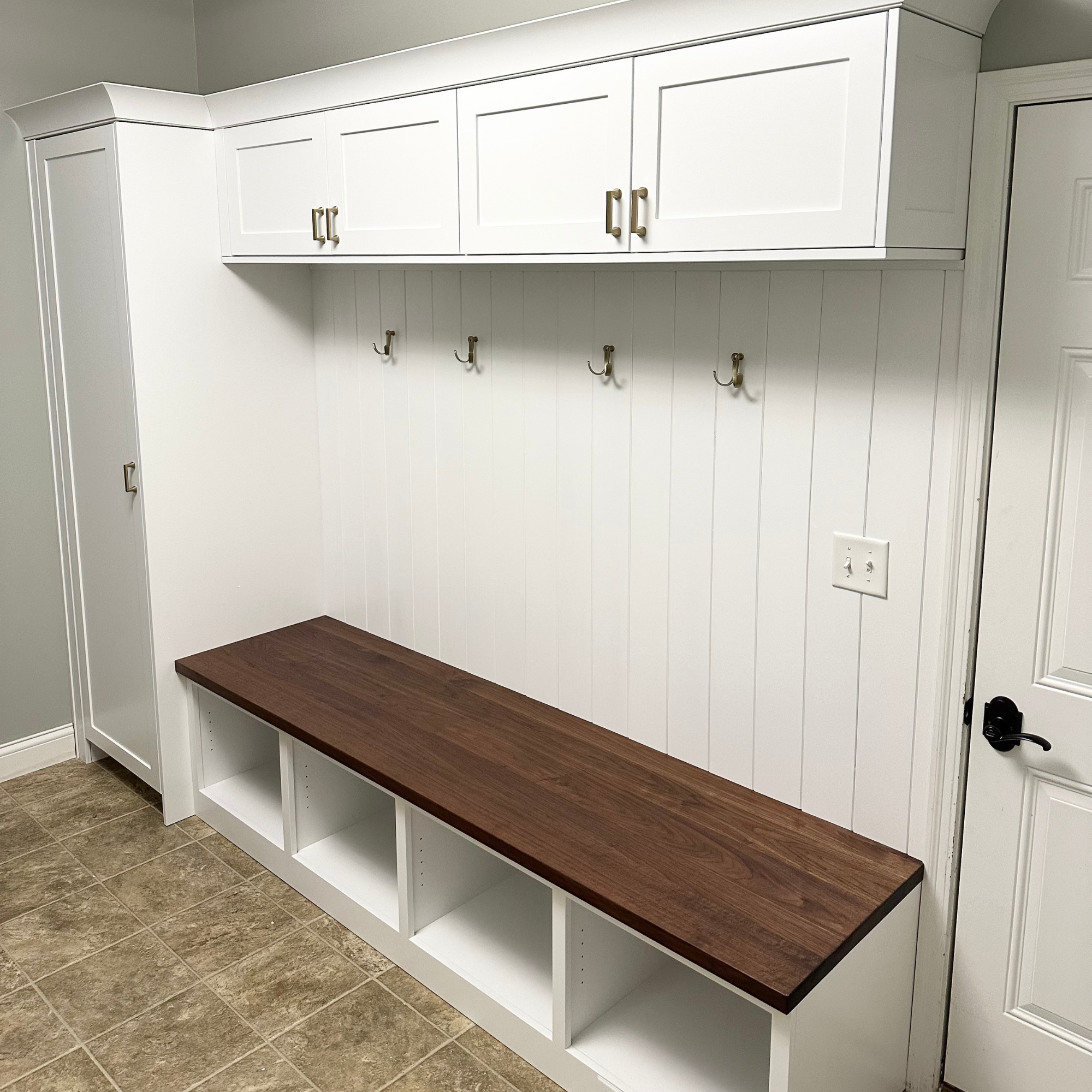 Custom white shaker-style mudroom bench unit featuring a dark stained walnut wood seat, open lower shoe cubbies, a vertical storage locker, brushed gold hardware handles, and a shiplap accent wall with wall hooks.
