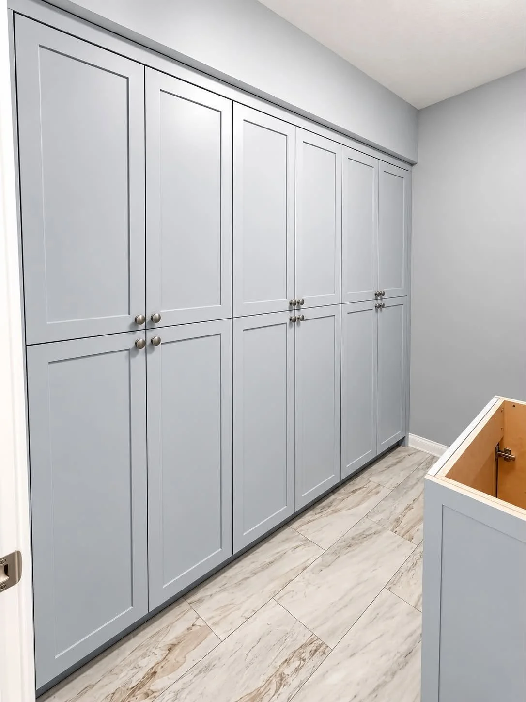 Closet with light grey cabinets and small round silver knobs, with light-colored tile flooring and a corner of an open cabinet drawer in a room with light grey walls.
